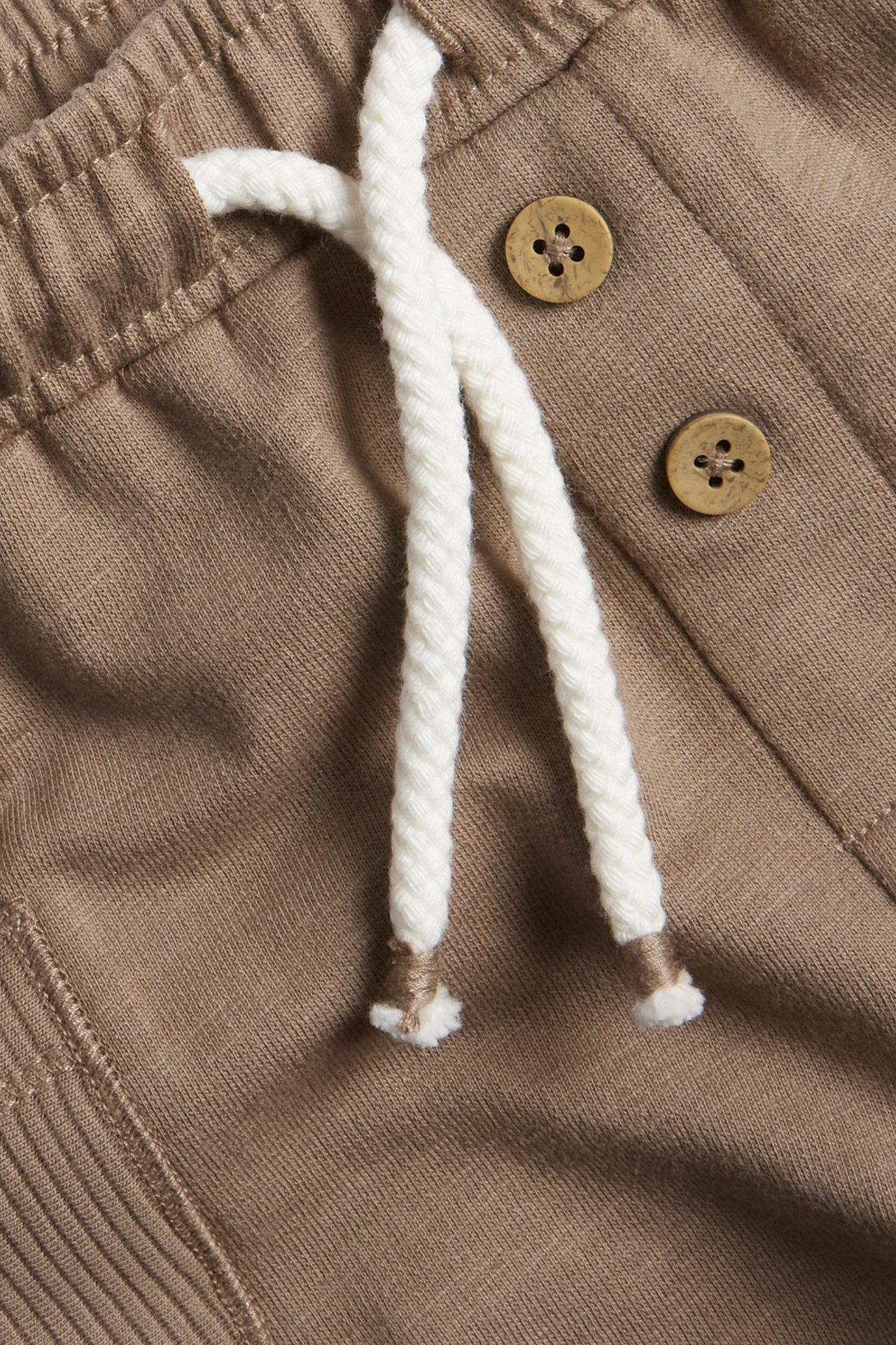 Nærbilde av brune slub-jersey shorts med elastisk midje, hvit snøring og dekorative knapper.