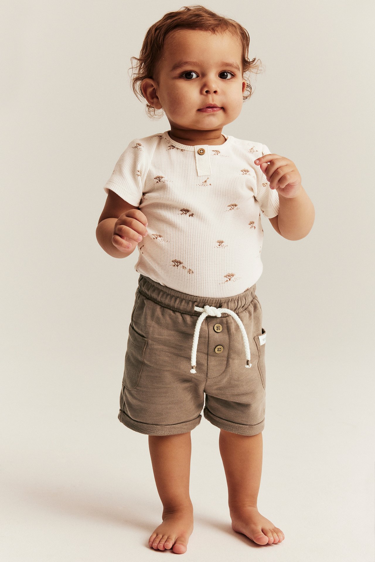 Frontbilde av en baby i brune bomullsshorts med hvit snøring og en hvitmønstret body.