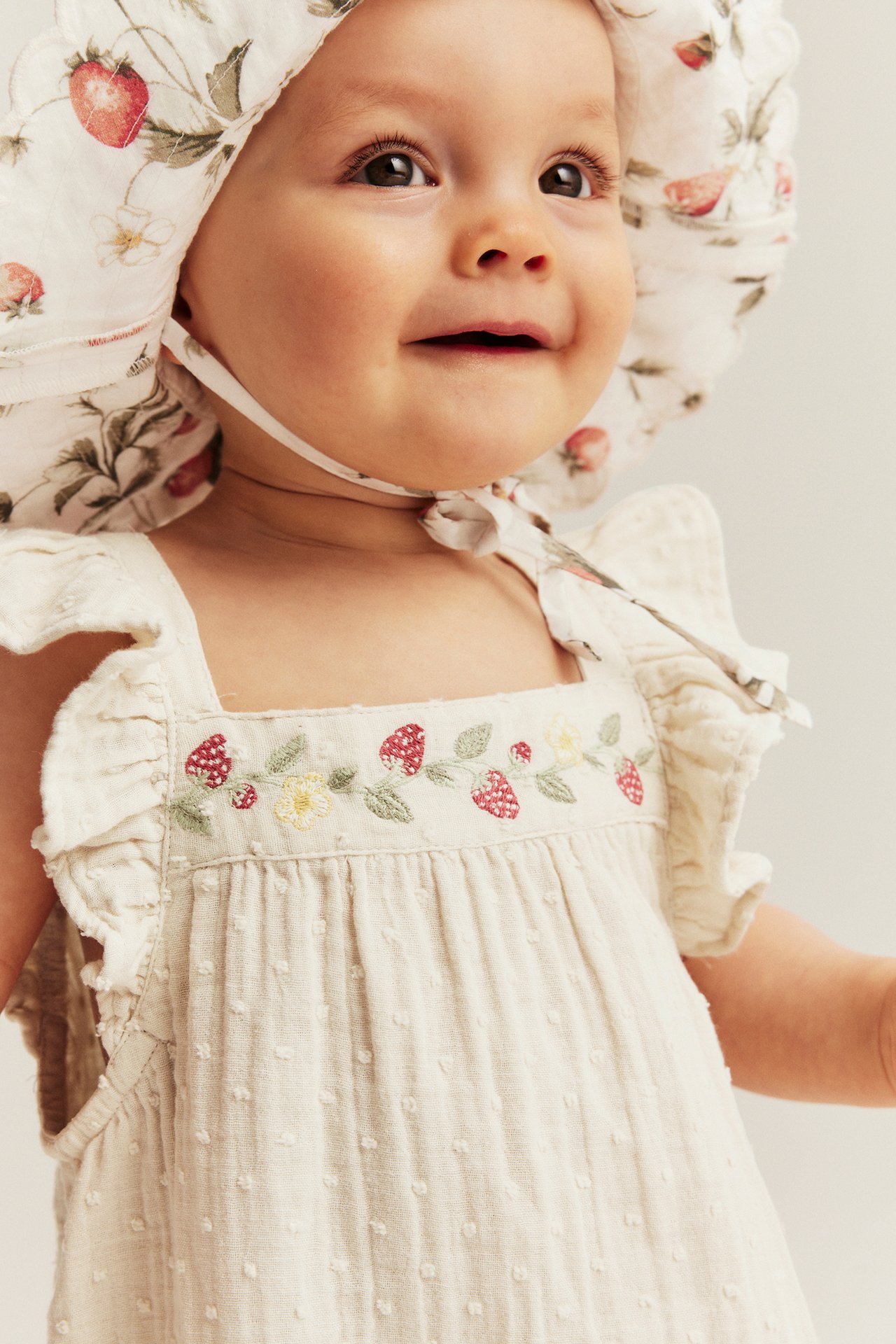 Zbliżenie na dziecko w jasnobeżowym tkanym kombinezonie z haftem truskawkowym i kwiecistym czepku.