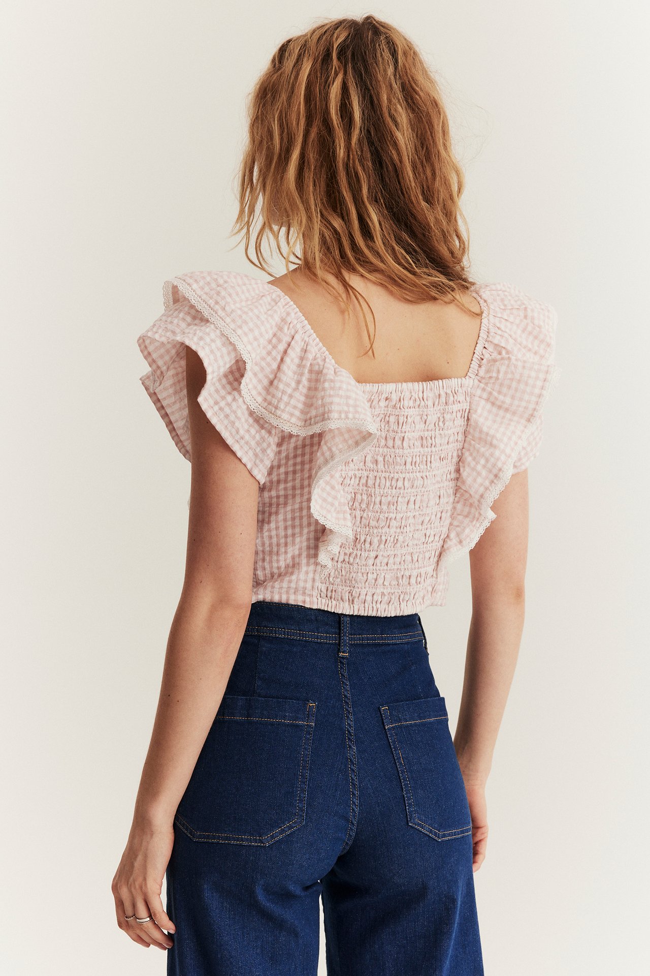 Bakifrån: kvinna i ljusrosa rutig blus med smock baktill, volangärmar och mörkblå jeans.