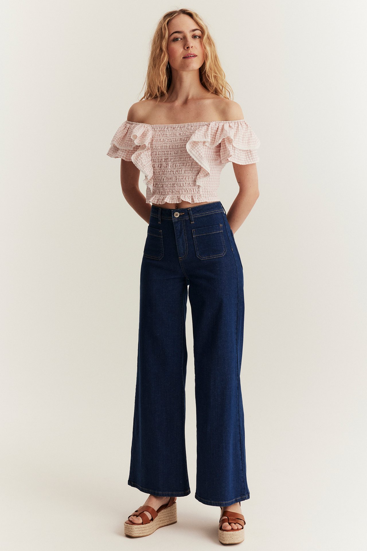Framifrån: kvinna i ljusrosa rutig smockad off-shoulder blus, mörkblå vida jeans, espadrillsandaler.