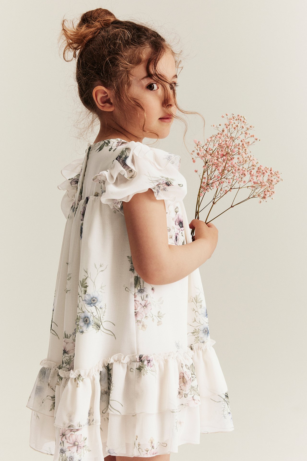 Sidebilde av et barn i offwhite blomstret chiffonkjole med volanger, holder rosa blomster.