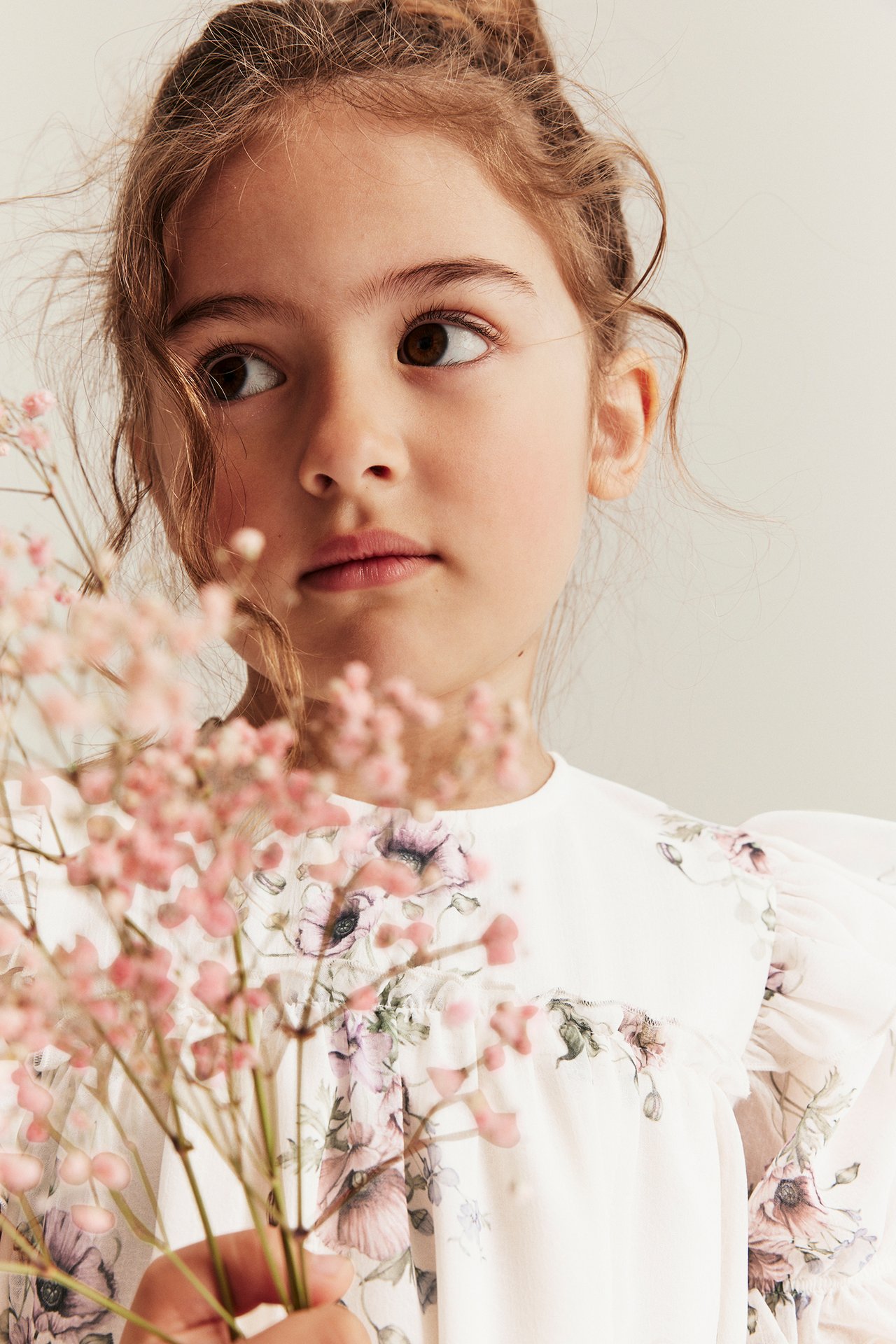 Nærbilde av en jente i offwhite blomstret chiffonkjole med puffermer, holder rosa blomster.