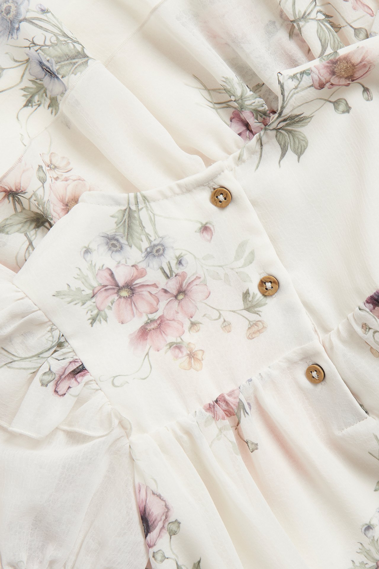 Nærbilde av en offwhite blomstret chiffonkjole med rosa og lilla blomster, rysjer og brune knapper.