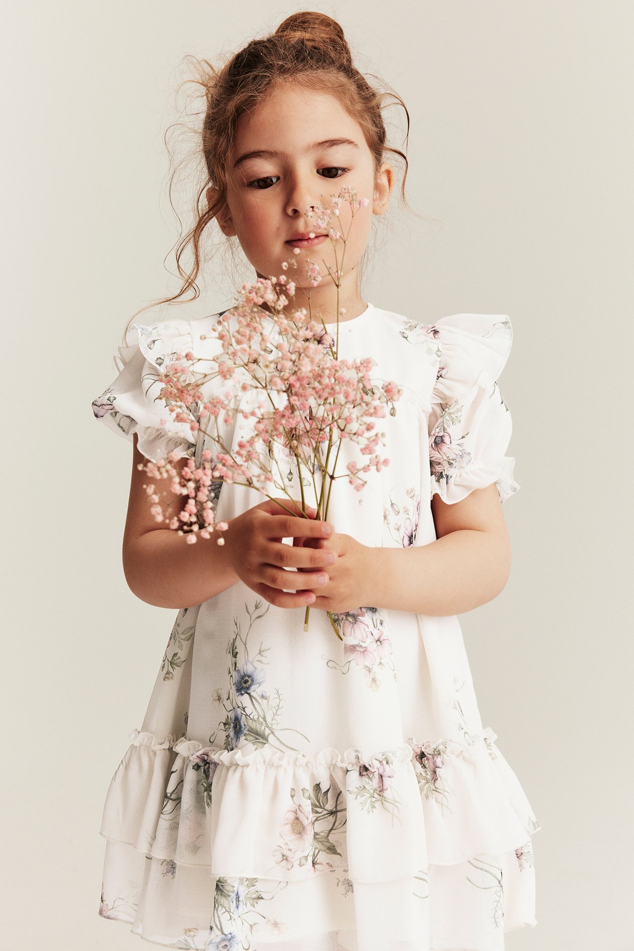Forfra, barn i offwhite blomstret chiffonkjole med volanger, holder rosa blomster.