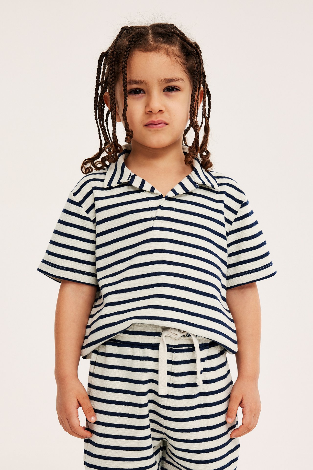 Frontbilde av et barn i lysebeige stripete bomullsshorts med snøring og matchende pikétrøye.