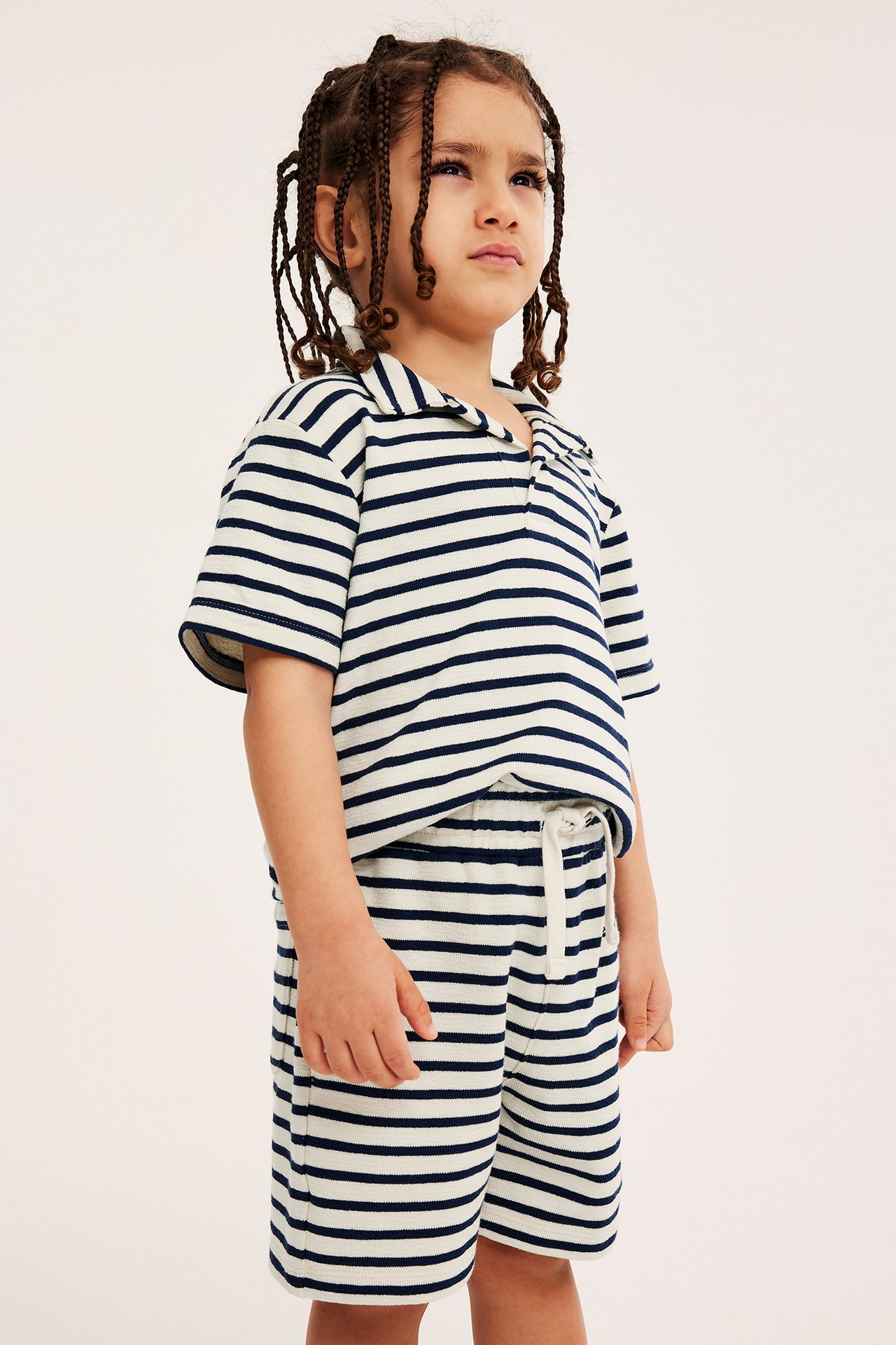 Frontbilde av et barn i lysebeige og marineblå stripete shorts med snøring og matchende piké t-skjorte.