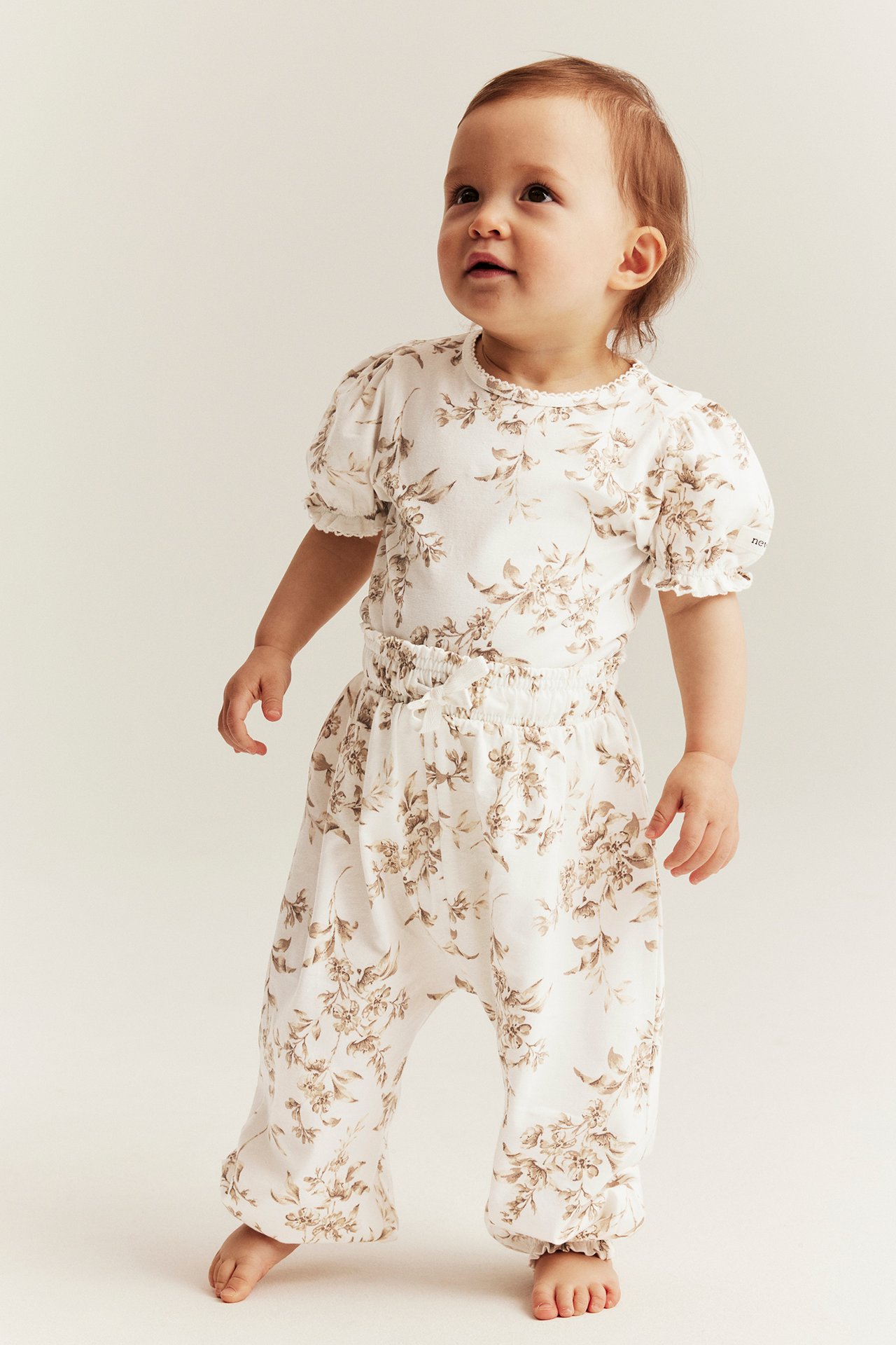 Framifrån: baby i offwhite blommigt jerseyset, topp med puffärm och vida byxor.
