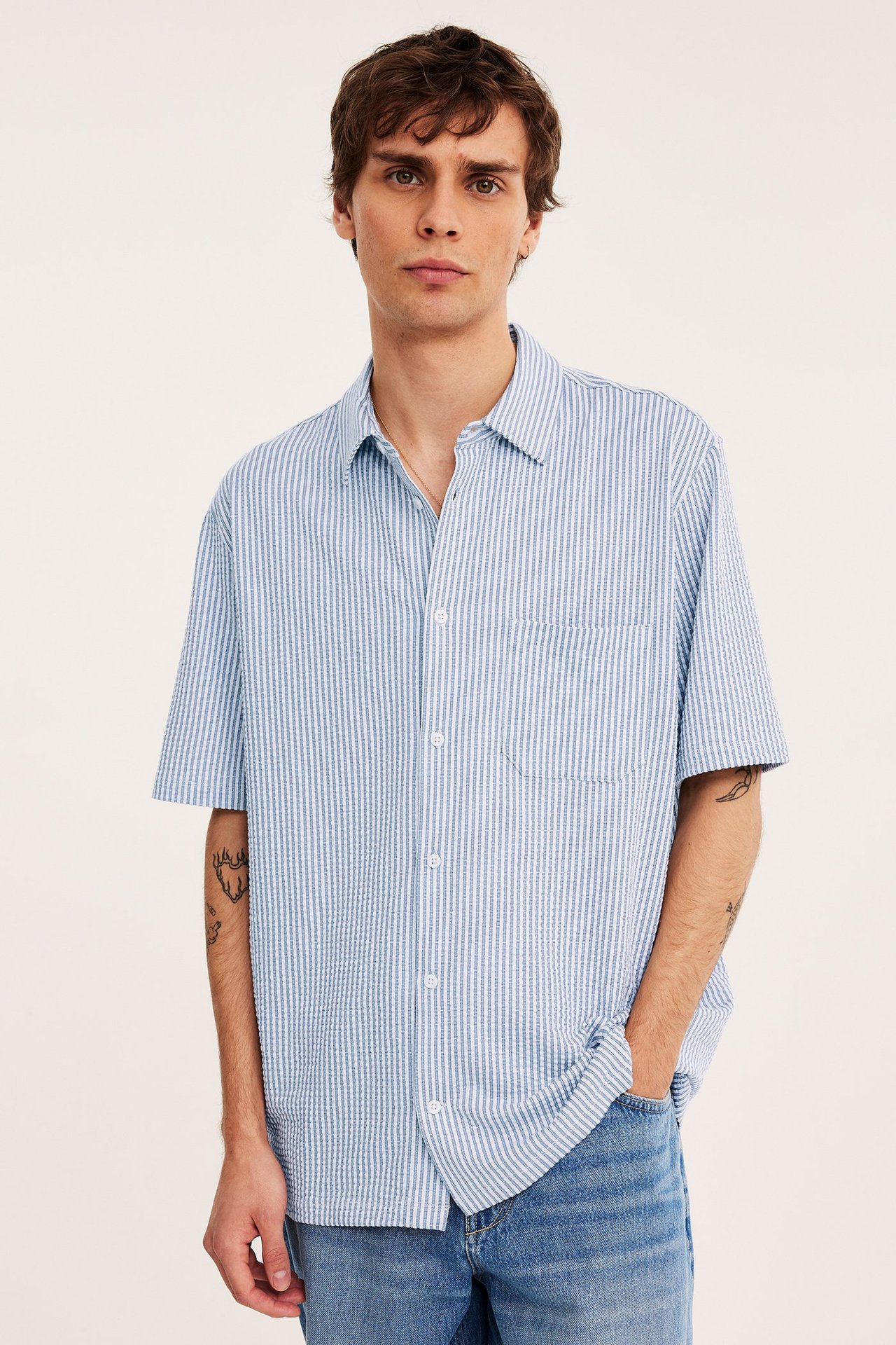 Edestäpäin mies yllään vaaleansinivalkoraidallinen lyhythihainen trikoopaita rintataskulla ja siniset farkut.