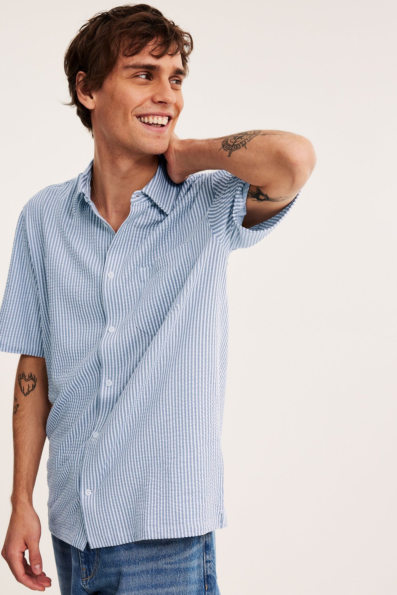 Edestä: Hymyilevä mies yllään vaaleansiniraidallinen lyhythihainen seersucker-paita rintataskulla ja siniset farkut.