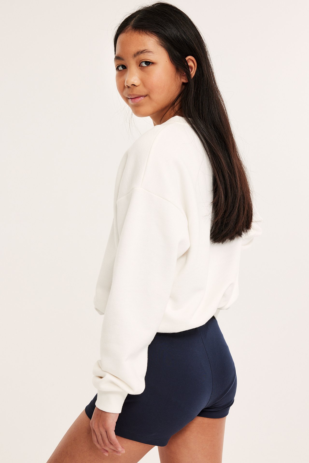 Sidovy av ett barn i mörkblå tajta cykelshorts och en krämvit långärmad sweatshirt.