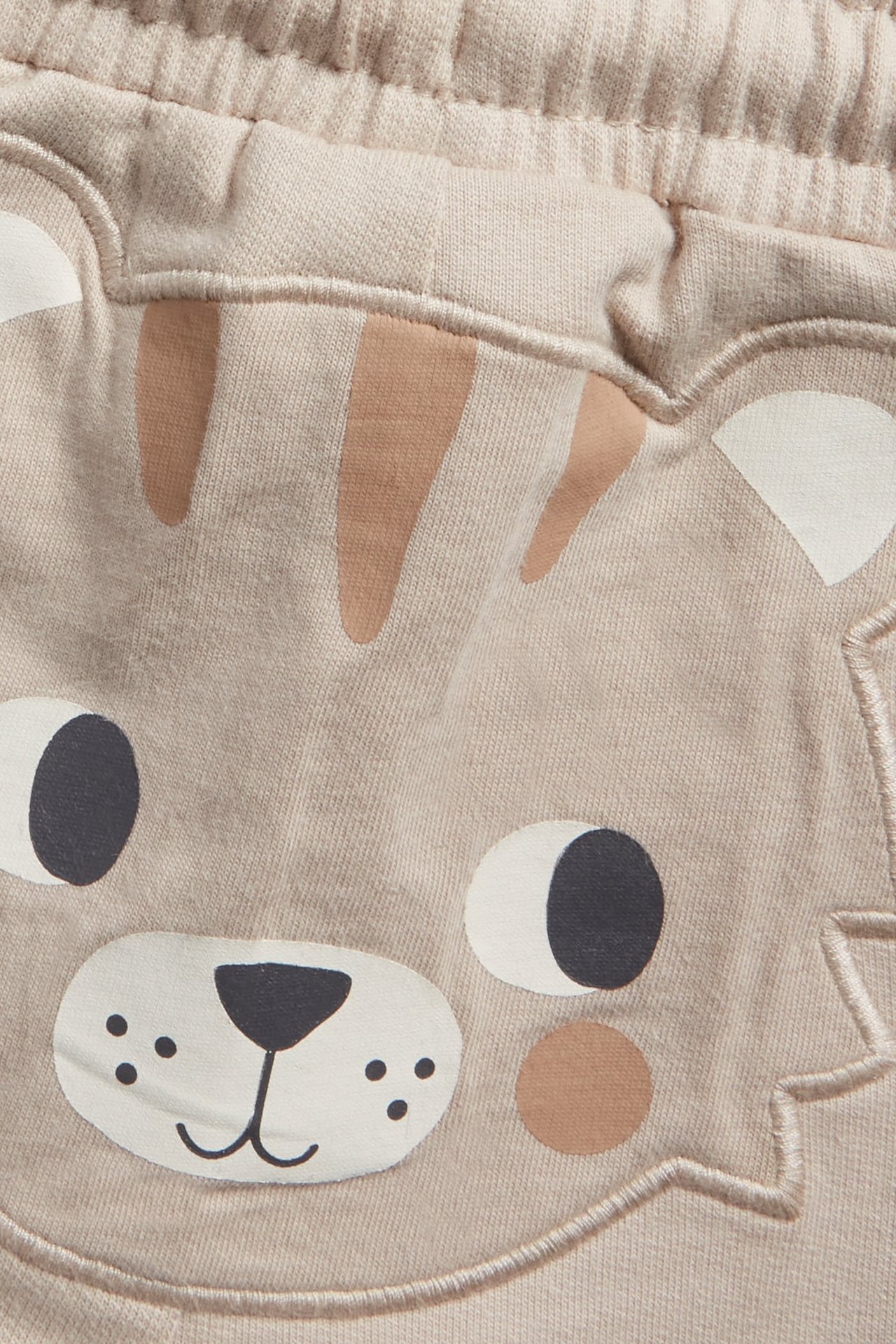 Nærbilde av beige baby-bomullsshorts med løveansikt-applikasjon og elastisk linning.