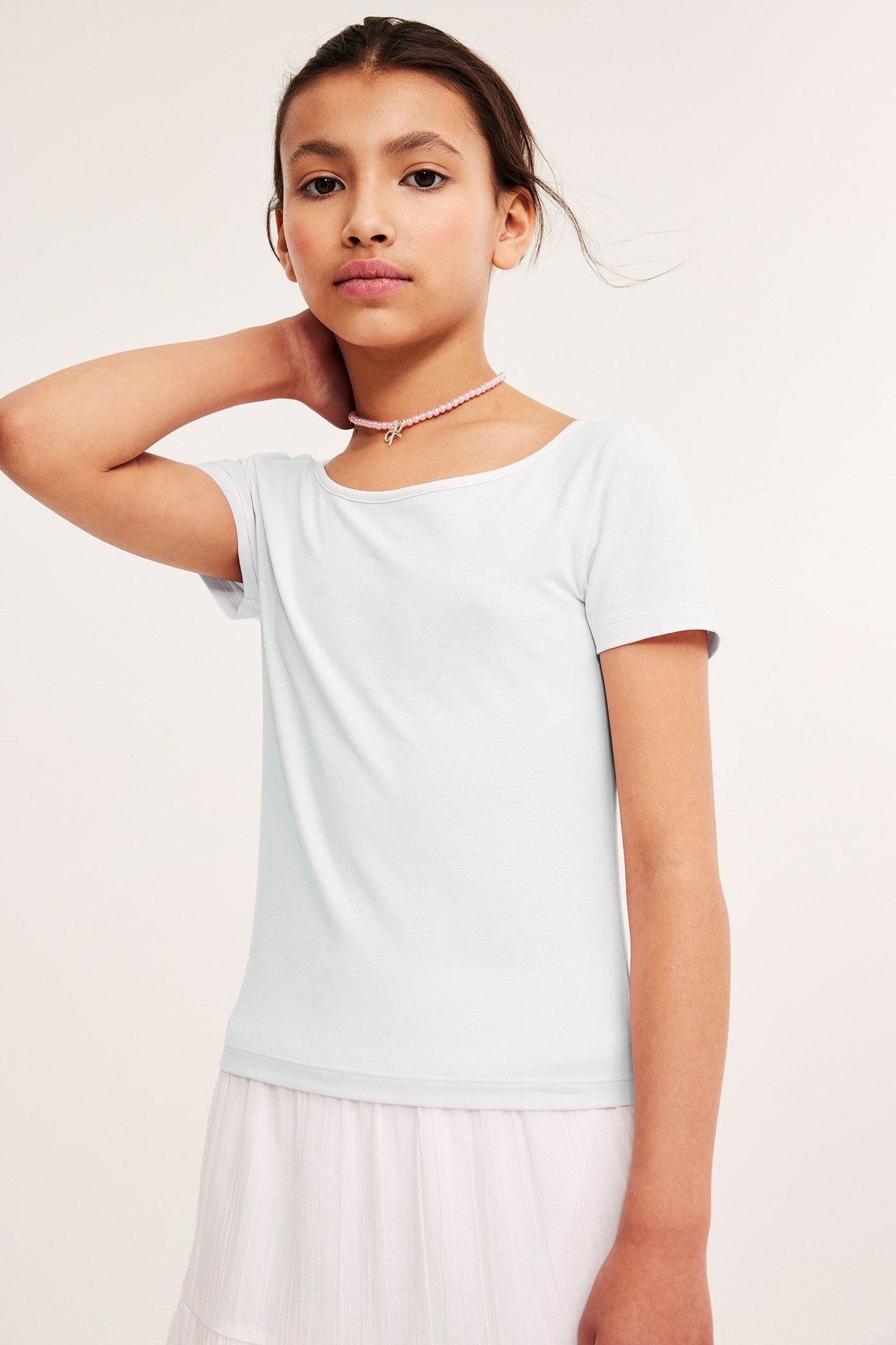 Framsida av ett barn i en vit åtsittande kortärmad topp, ljusrosa kjol och rosa pärlhalsband.