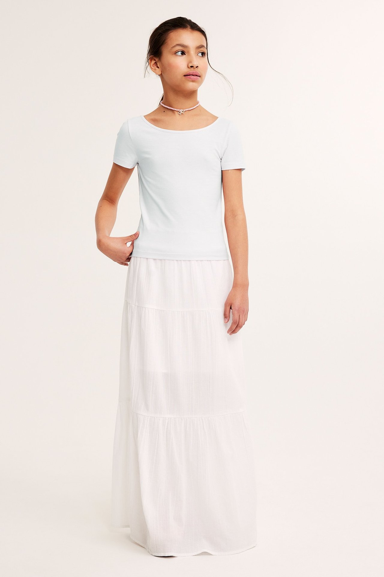 Frontvy av ett barn i en vit åtsittande kortärmad topp, en vit volangmaxikjol och ett rosa pärlhalsband.