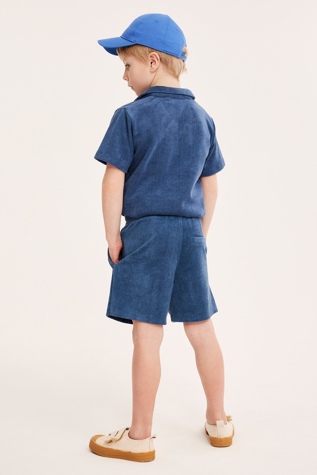 Ryggvisning av et barn i mørkeblå frottéshorts, matchende pikétrøye, blå caps og beige sneakers.