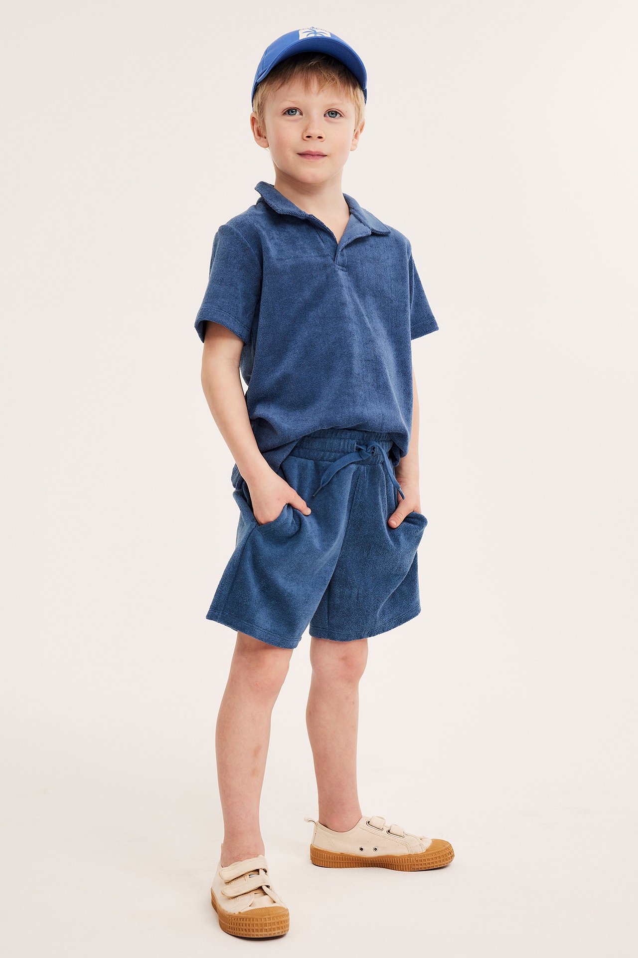 Forfra, et barn i mørkeblå frottéshorts, matchende pikétrøye, blå caps og beige sko.