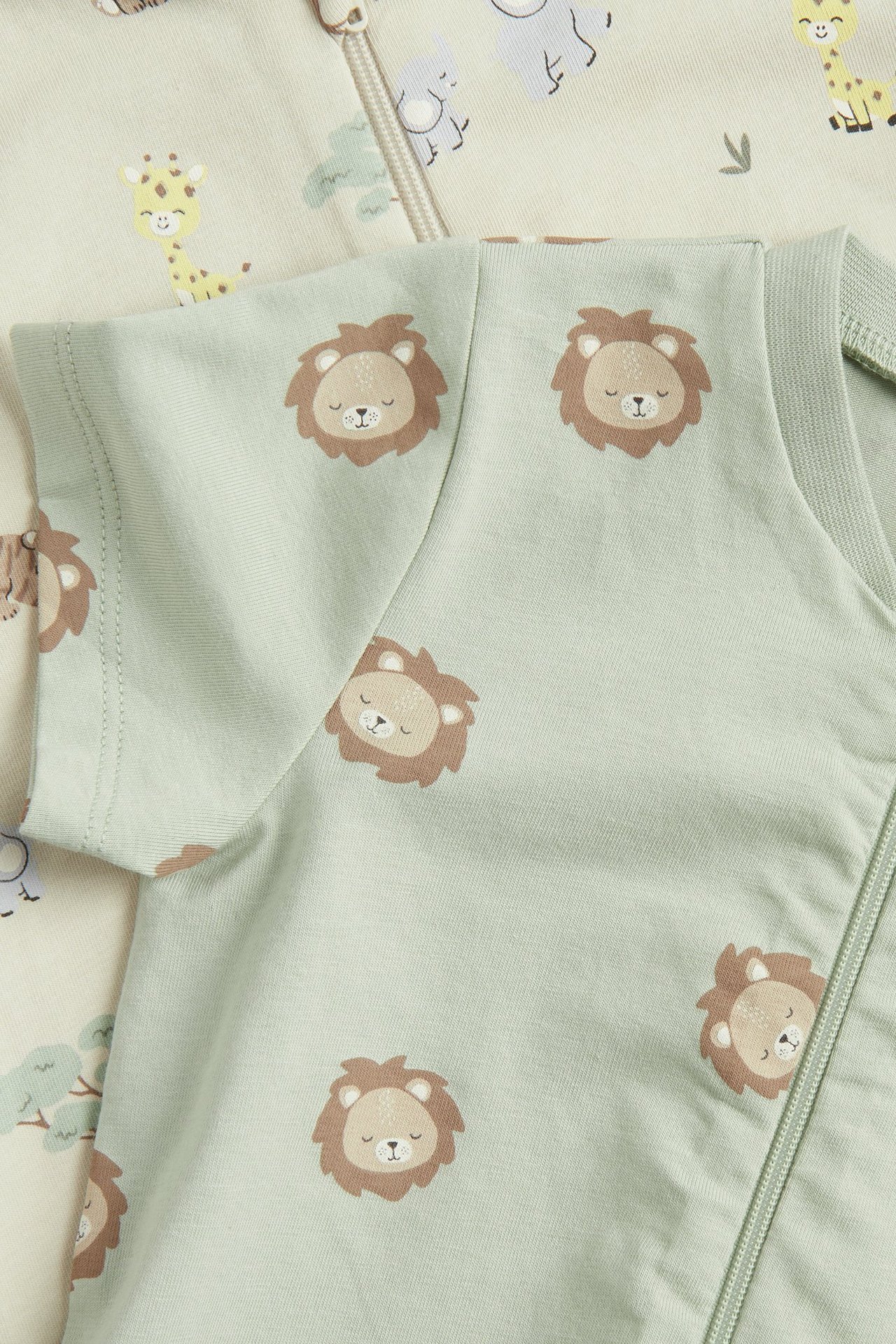 Zbliżenie na jasnozielone body niemowlęce z krótkim rękawem w lwy, obok beżowe body w zwierzęta.