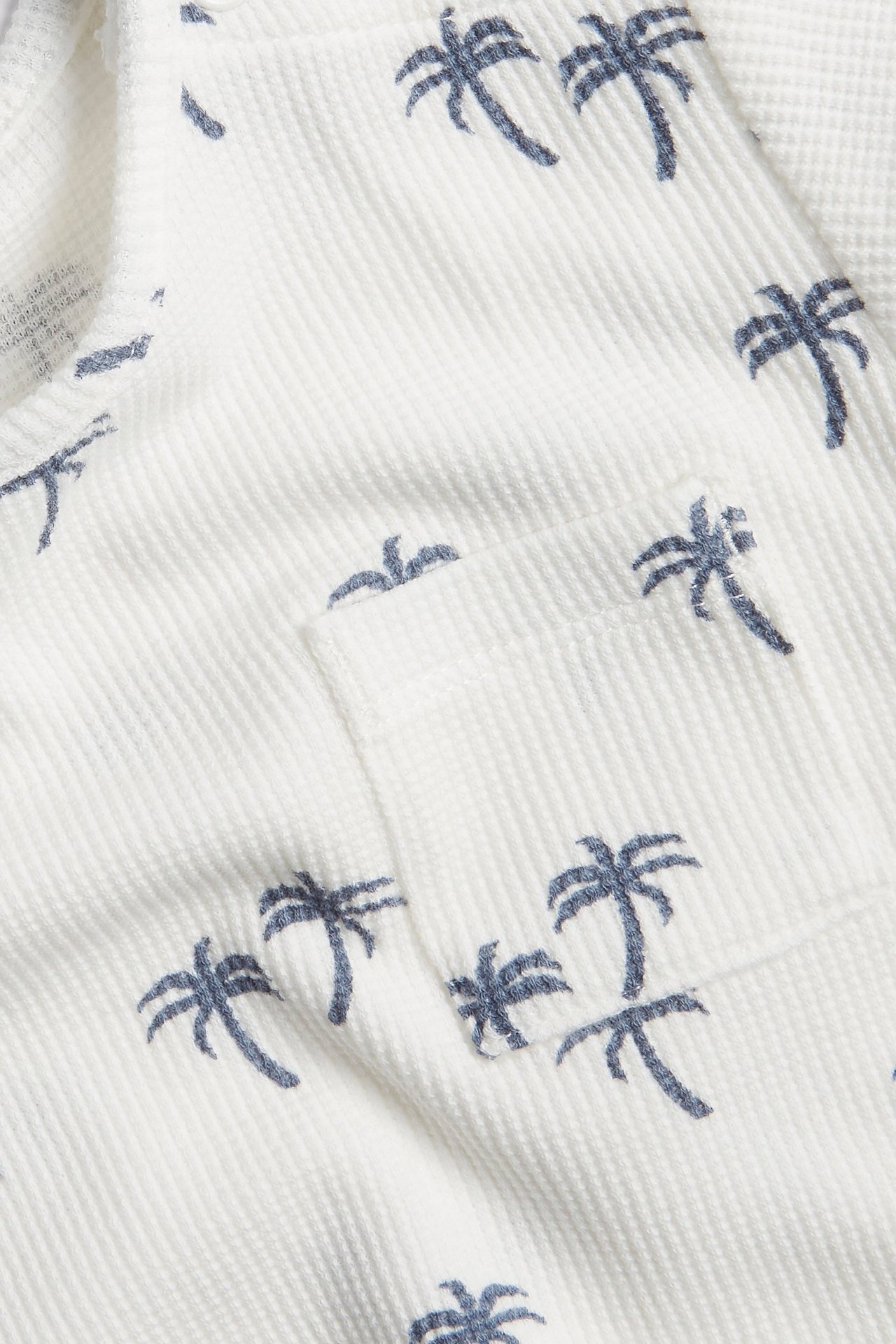 Zbliżenie: Biały t-shirt niemowlęcy z waflowej dzianiny z niebieskim wzorem palm i kieszenią.