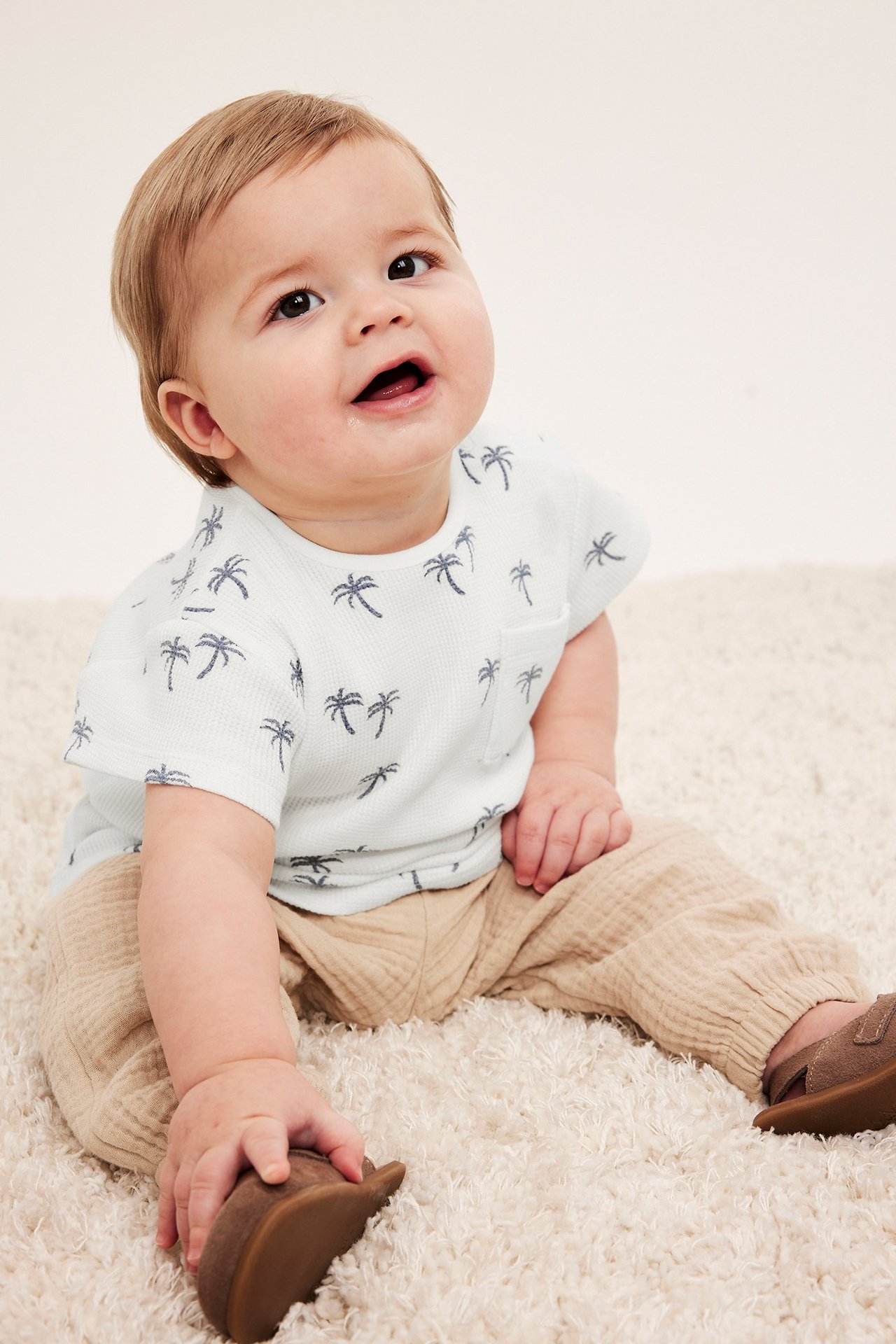 Widok z przodu: niemowlę w ecru waflowym T-shircie w palmy, beżowych spodniach i brązowych butach.