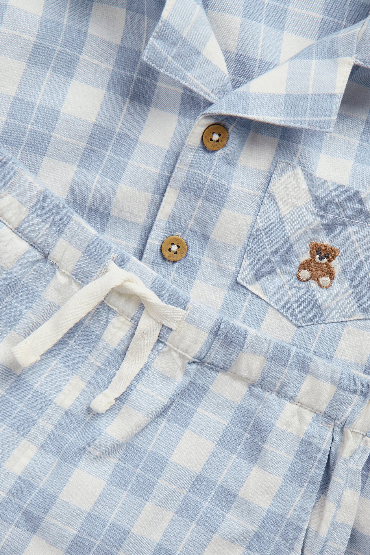 Närbild av ett blåvitt rutigt pyjamasset. Skjortan har knappar, nallebroderi på fickan och dragsko i shortsen.