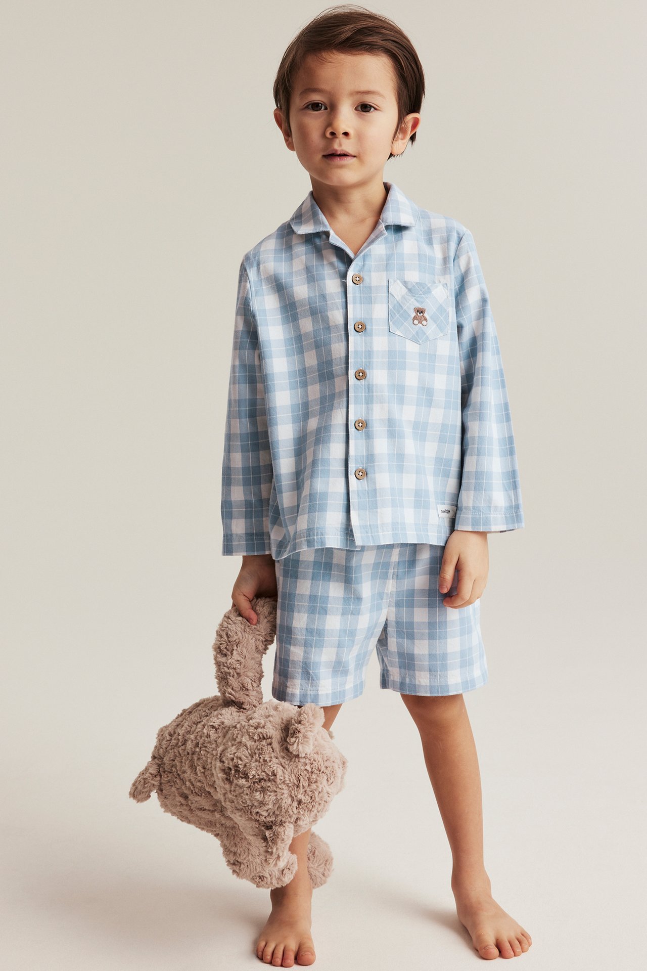 Framifrån, ett barn i blåvitrutig pyjamas i bomull, håller en brun fluffig nallebjörn.