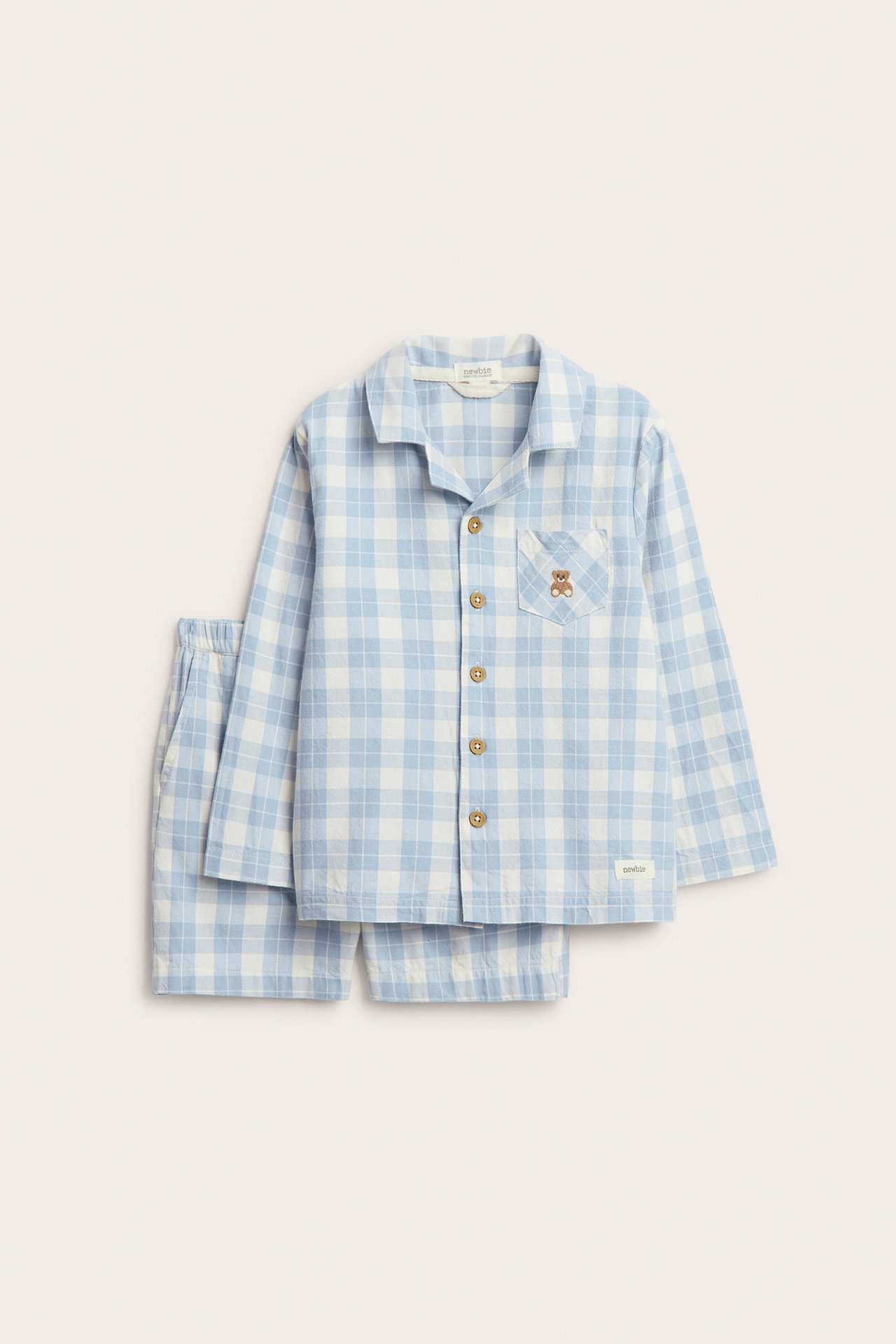 Framsida av en rutig pyjamas i blå och vit bomull för barn: långärmad skjorta med nallebroderi, shorts.