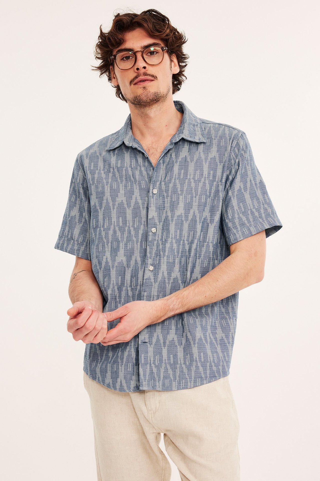 Edestä kuvattu mies tummansinisessä kuviollisessa lyhythihaisessa puuvillapaidassa, vaaleanbeigeissä housuissa ja ruskeissa silmälaseissa.
