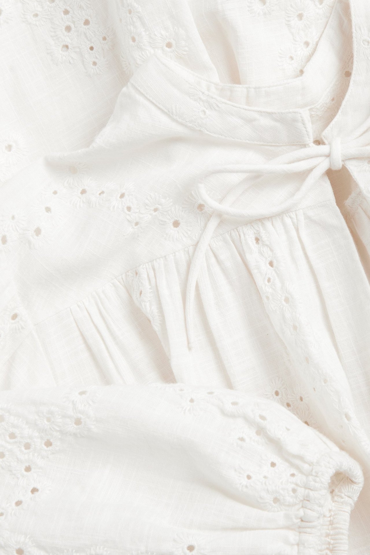 Zbliżenie białej bawełnianej sukienki z haftem angielskim, wiązaniem pod szyją i marszczonymi detalami.