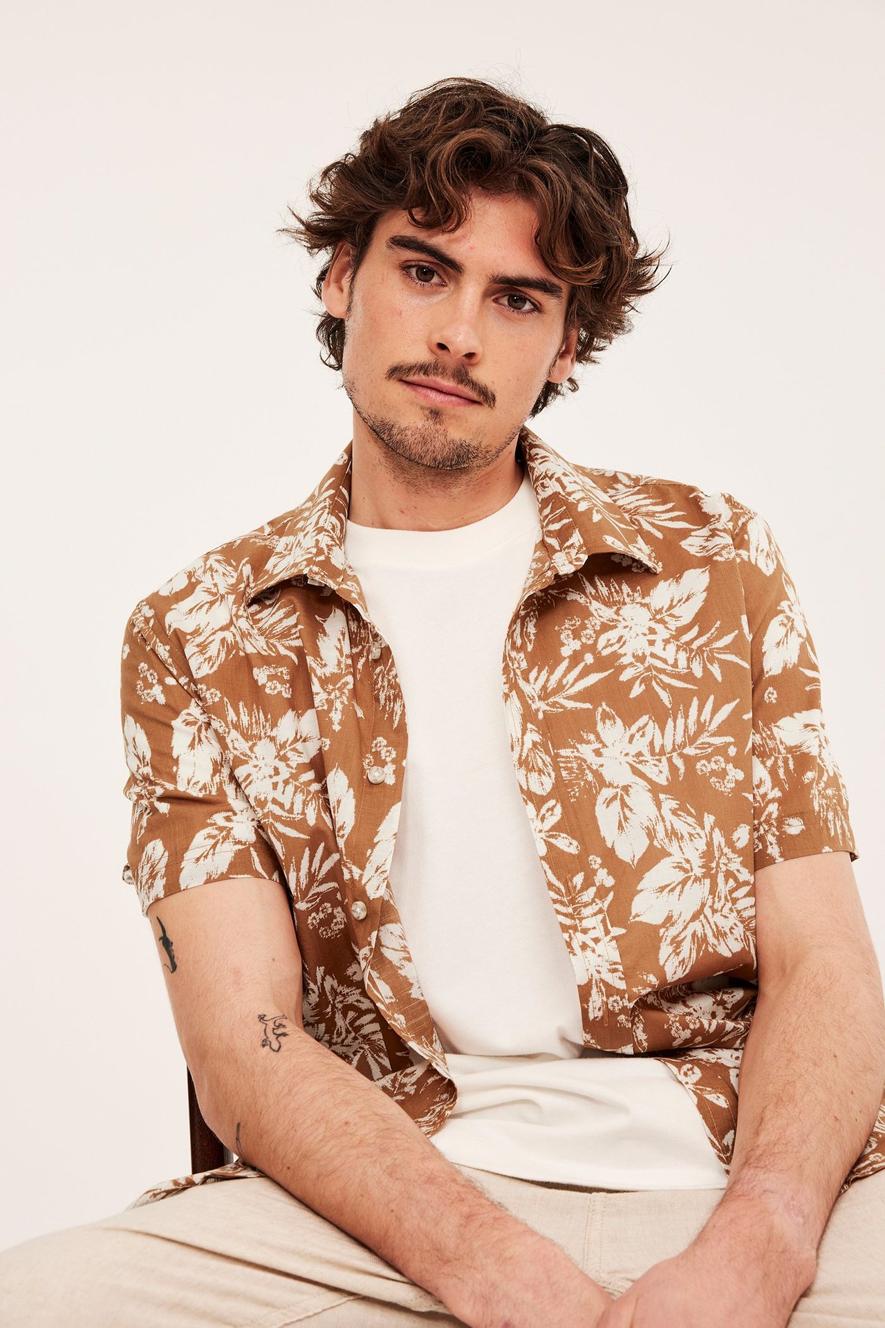 Edestäpäin mies avonaisessa ruskeassa lyhythihaisessa paidassa, jossa valkoinen kukkakuvio, valkoisen t-paidan päällä.