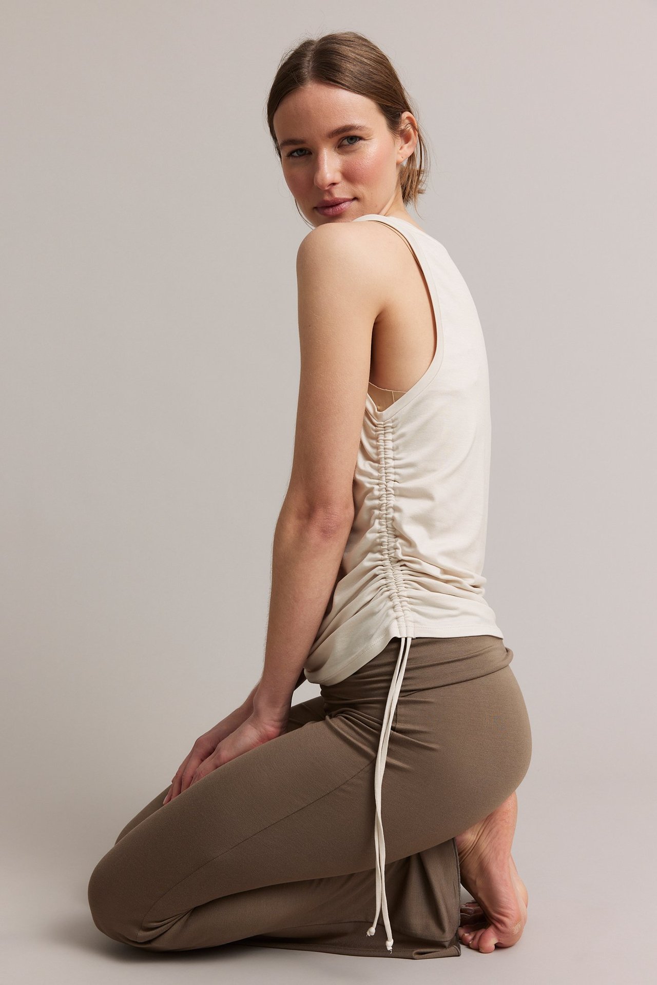 Sidevisning av kvinne som kneler, iført en lys beige tencel-topp med snøring i siden og brune bukser.