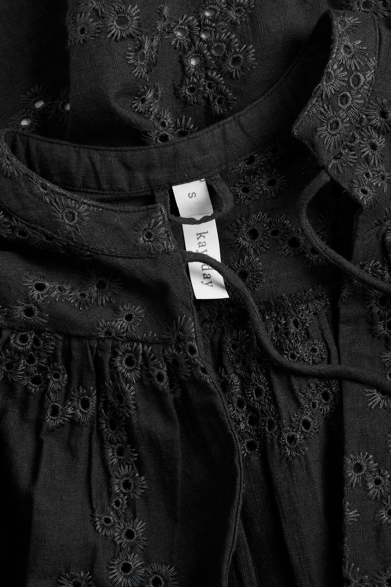 Nærbilde av en svart bomullsbluse med broderie anglaise, ståkrage og knytebånd.