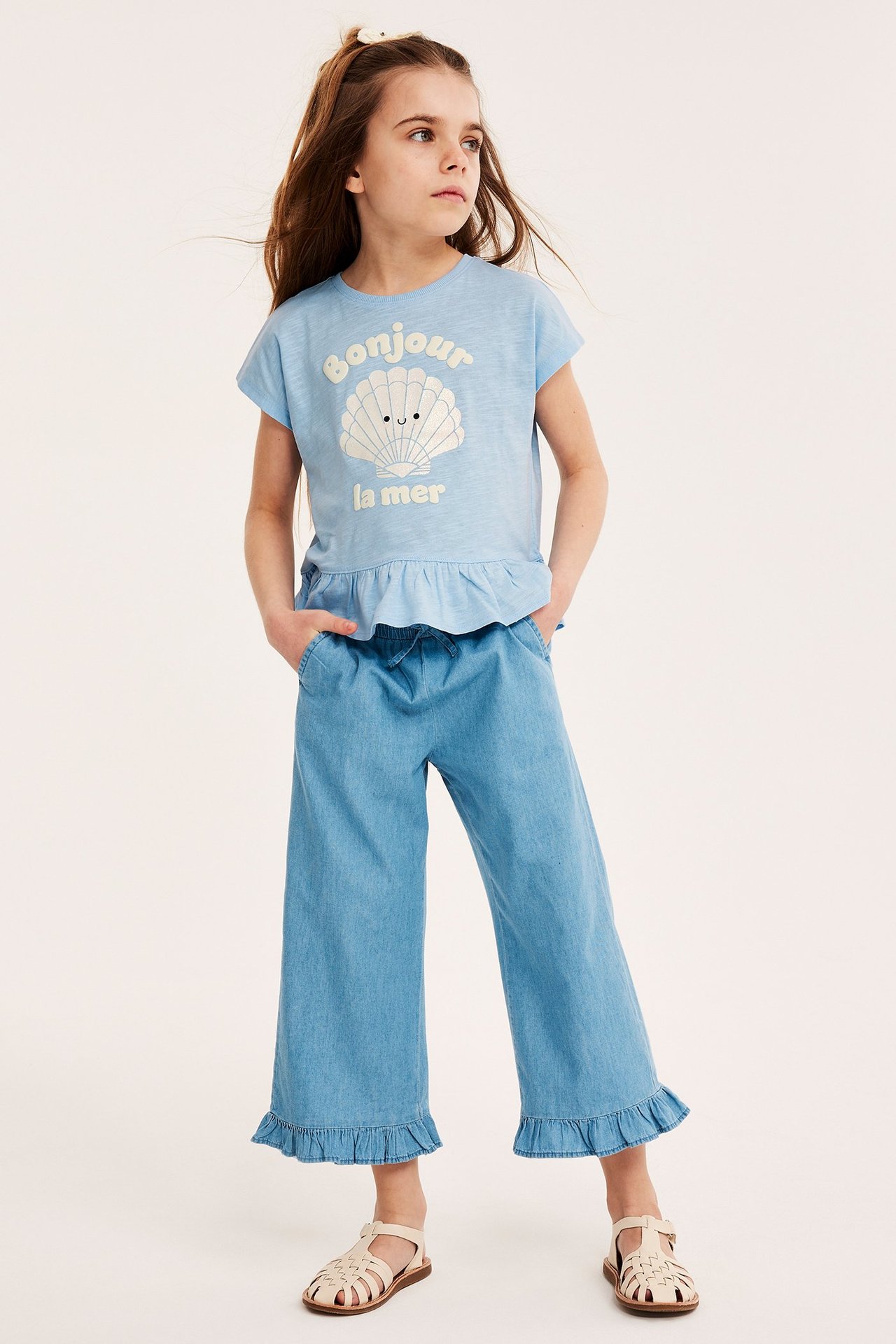 Widok z przodu: dziecko nosi niebieski bawełniany top z nadrukiem muszli, jasnoniebieskie spodnie i beżowe sandały.