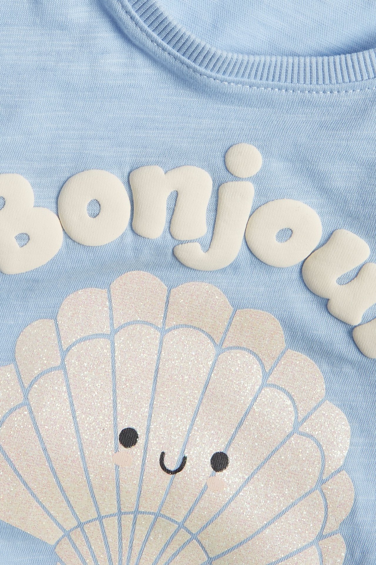Zbliżenie na jasnoniebieski bawełniany top dziecięcy z napisem 'bonjour' i brokatowym, uśmiechniętym nadrukiem muszli.