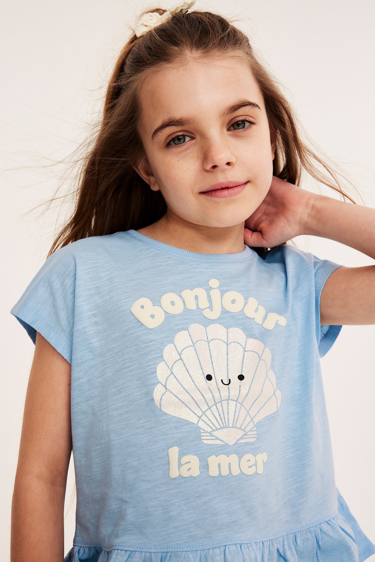 Widok z przodu: Dziecko w jasnoniebieskim topie bawełnianym z nadrukiem muszli, falbanką i kokardą.
