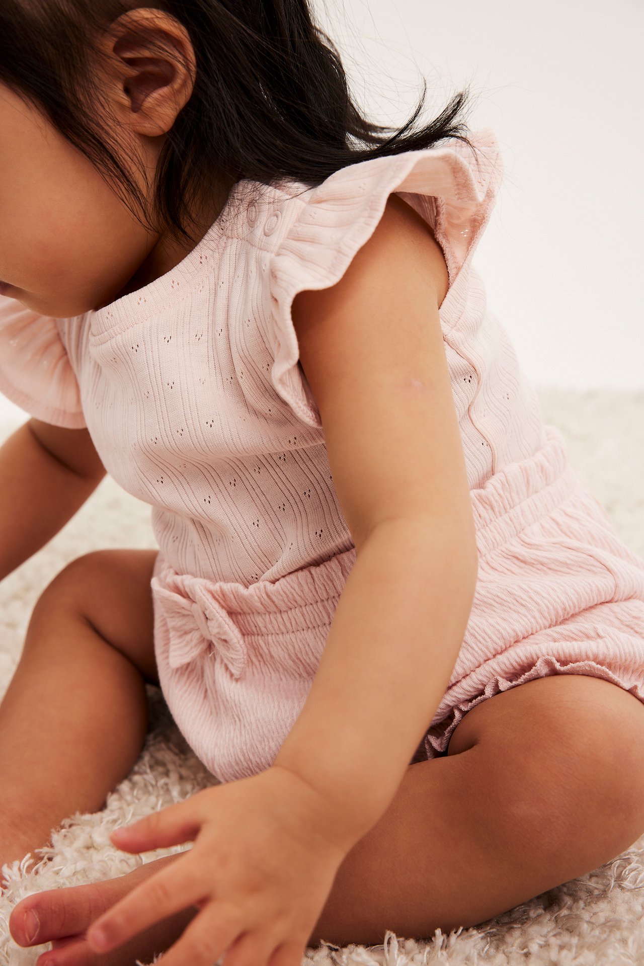 Nærbilde sidevisning av en baby i lys rosa krøllete shorts med sløyfe og ribbet topp.