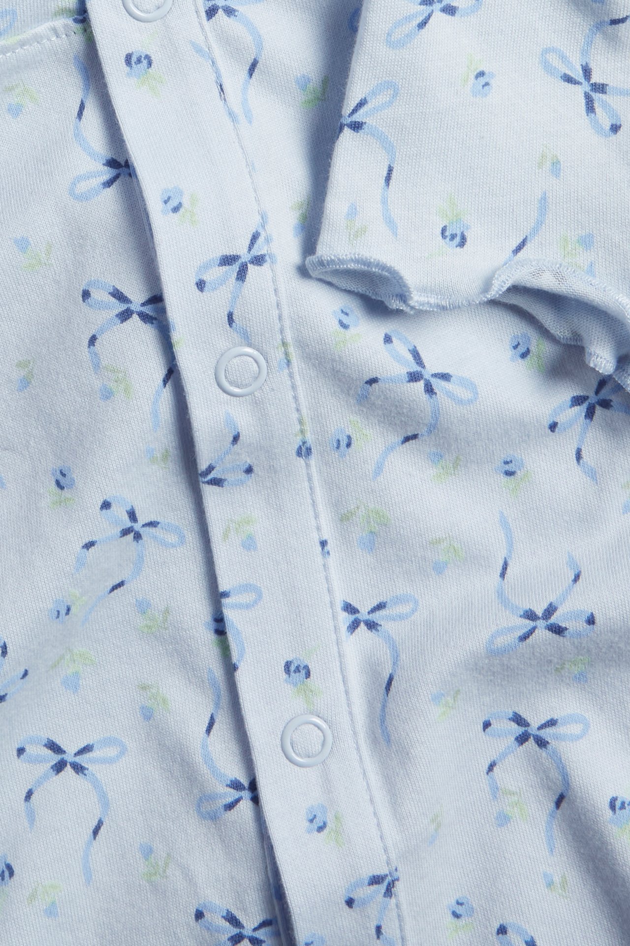 Närbild av en ljusblå pyjamas-jumpsuit i bomull med blå rosett- och blommönster, vita tryckknappar.
