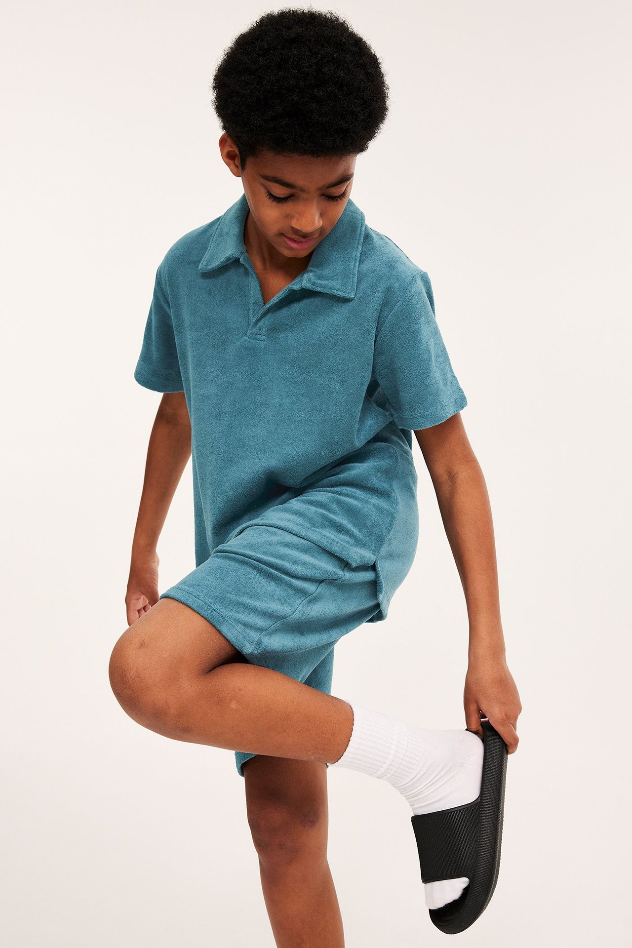 Widok z przodu dziecka w turkusowej koszulce polo z frotte i pasujących szortach, poprawiającego czarny klapek.