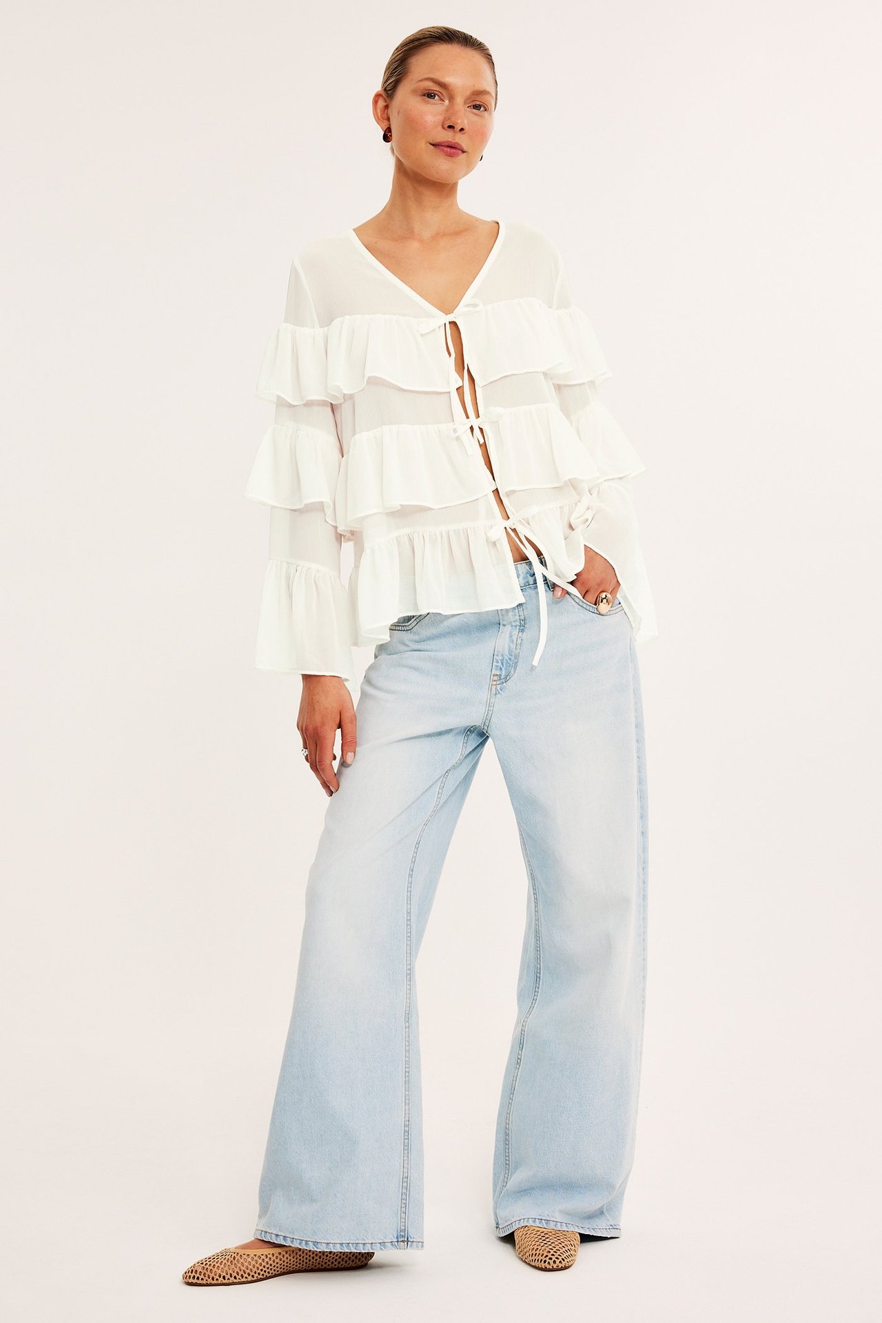 Framifrån: Kvinna i offwhite chiffongblus med volanger och knytband, ljusblå vida jeans, beiga vävda ballerinaskor.