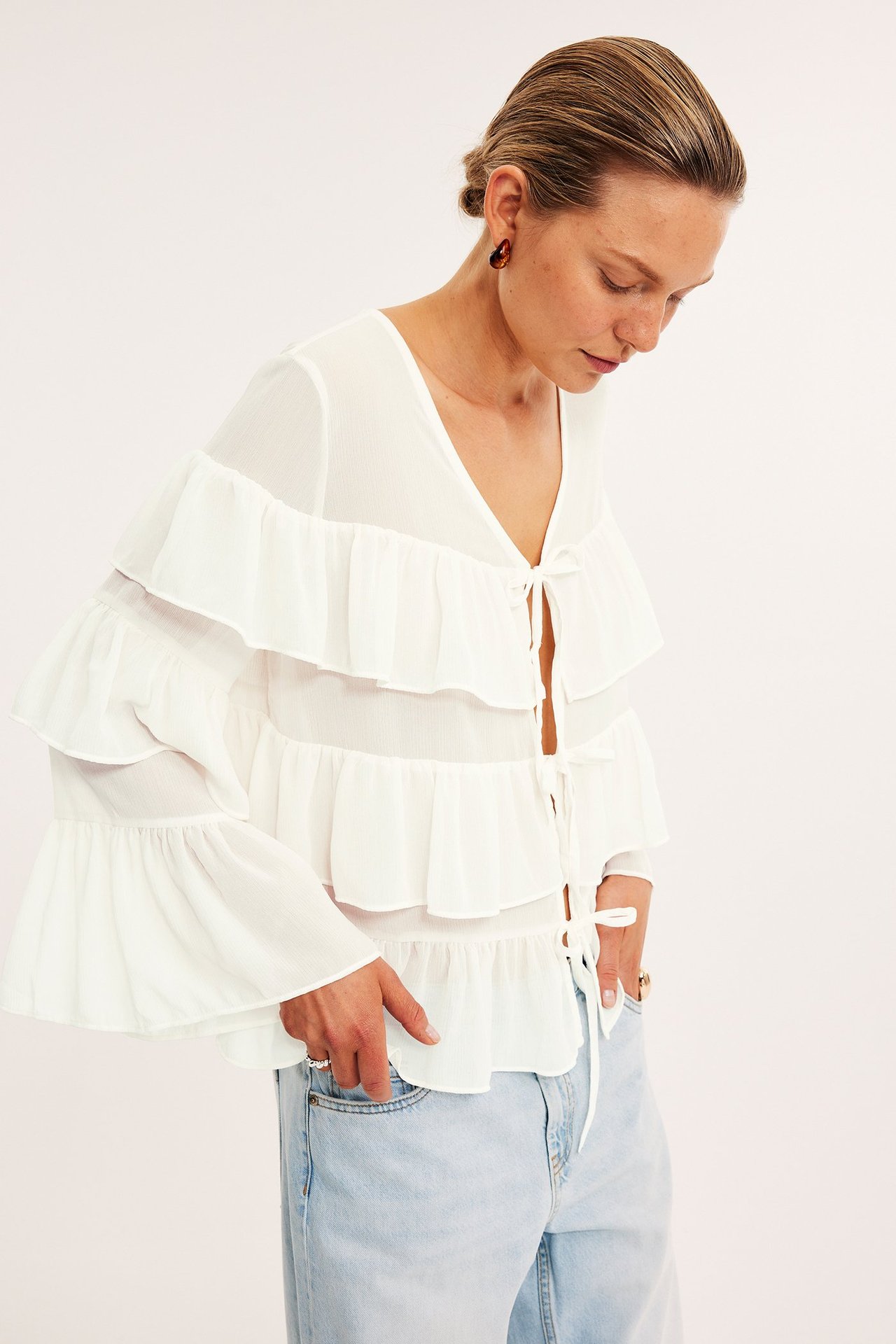 Frontvy av kvinna i offwhite chiffongblus med volanger och knytband, ljusblå jeans, bruna örhängen.