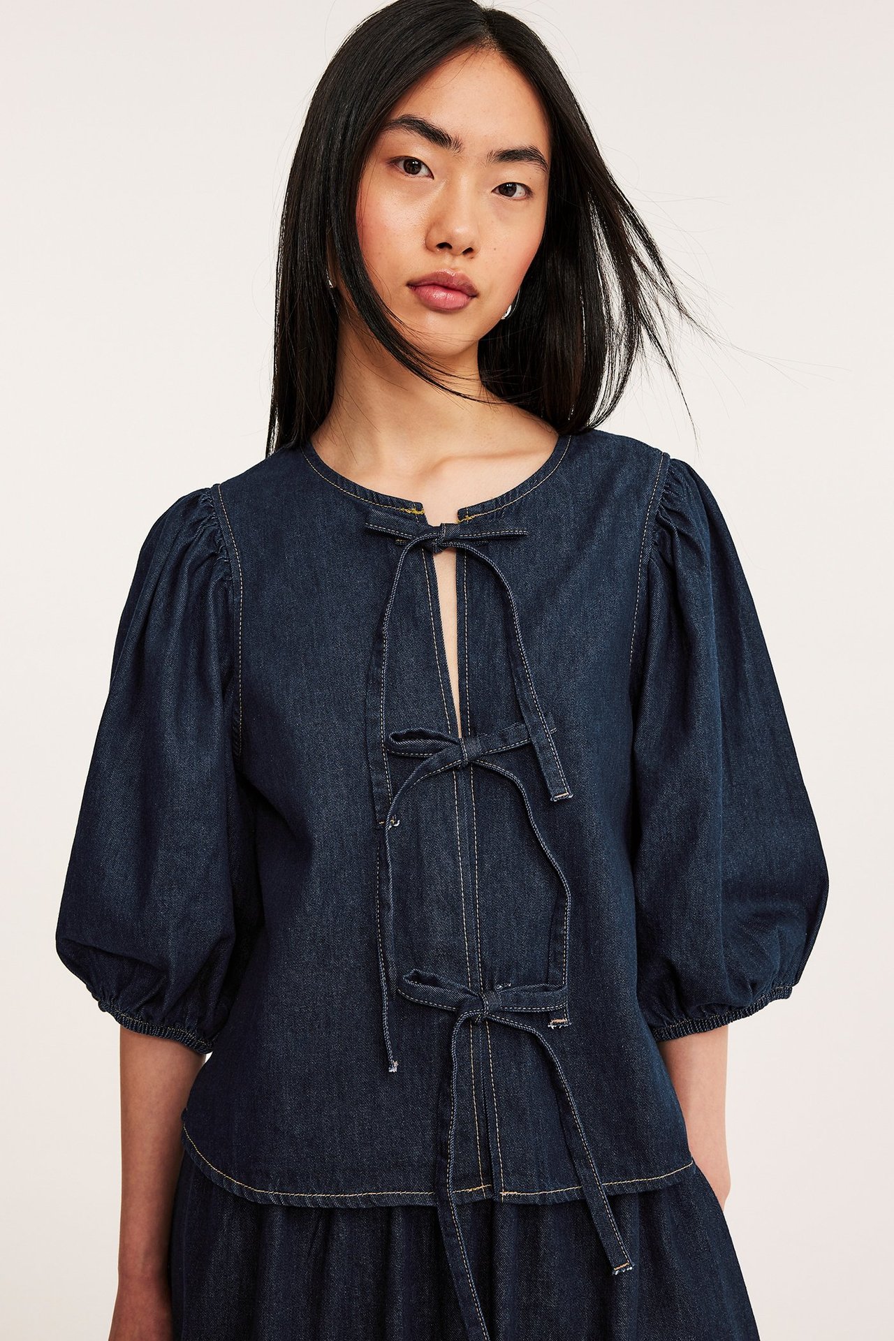Edestäpäin, nainen tummassa denim-puserossa, jossa edessä nauhat ja pussihihat, sekä yhteensopiva hame.