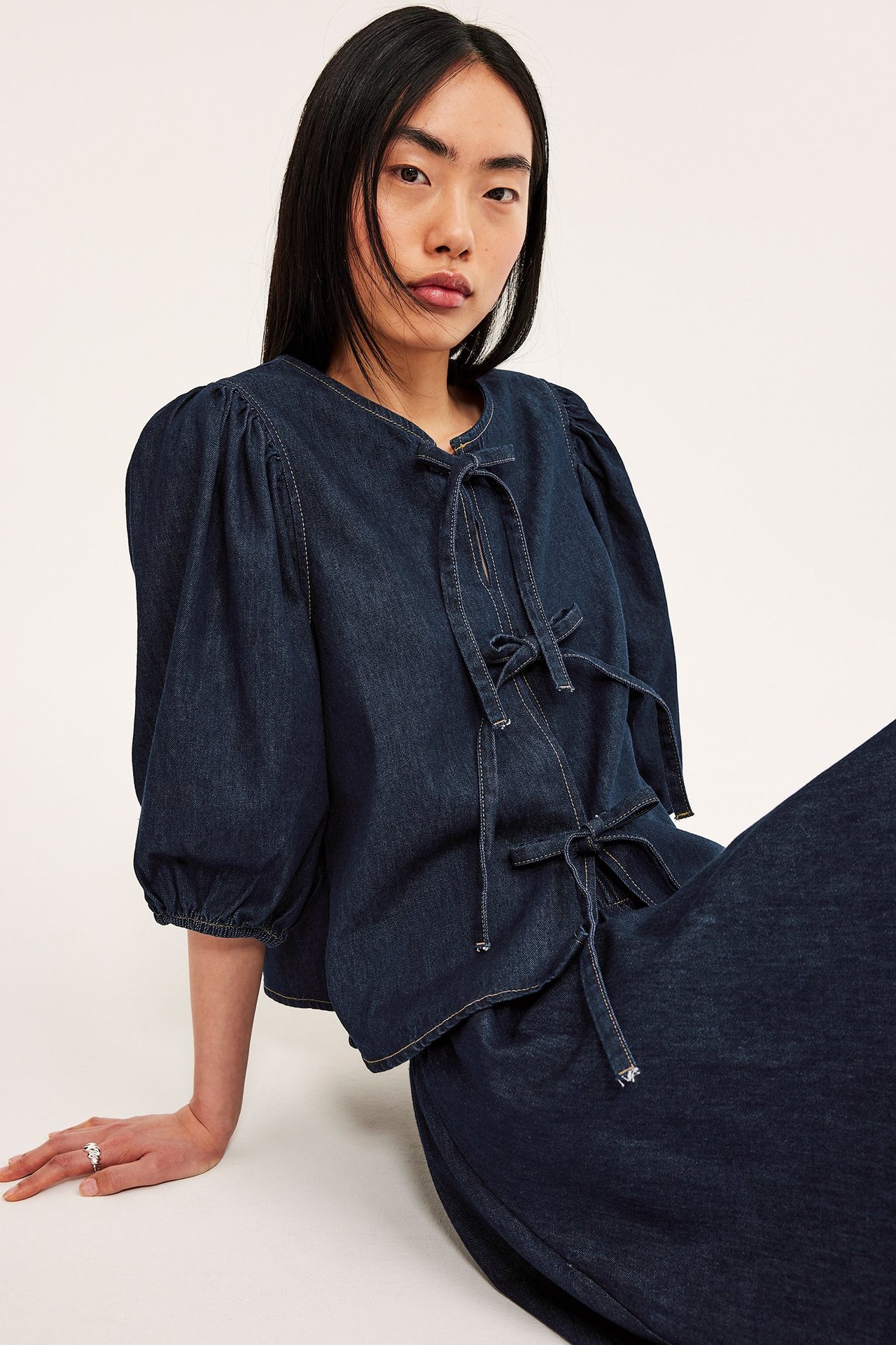 Widok z przodu: kobieta w ciemnej bluzce dżinsowej z bufiastymi rękawami i wiązaniami, z ciemnymi spodniami dżinsowymi.