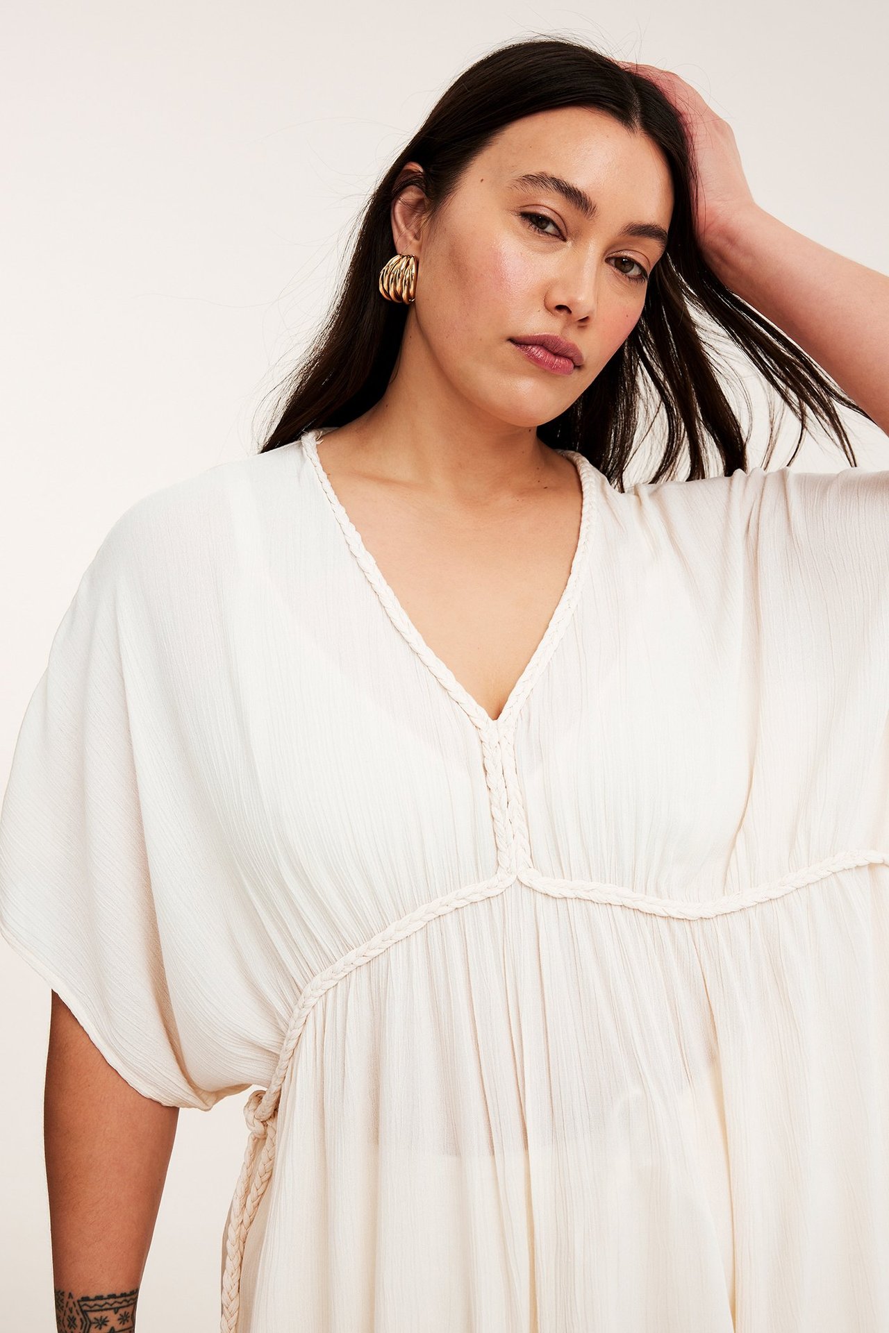 Nærbilde forfra av kvinne i offwhite krøllet viskosekjole med flettede detaljer og gullfargede øreringer.