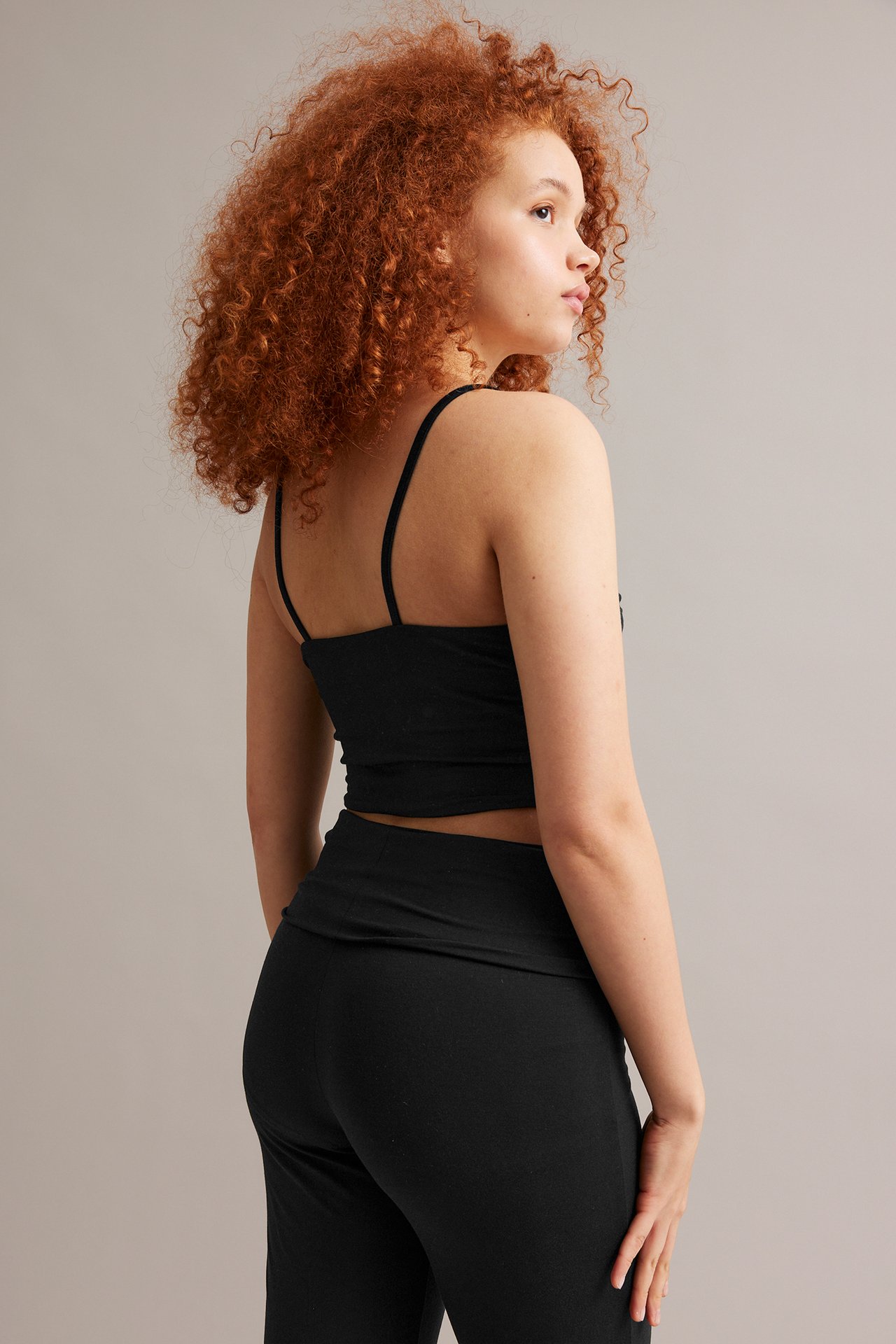 Rygg-/sidovy av kvinna i svart åtsittande kort linne och svarta högmidjade byxor.