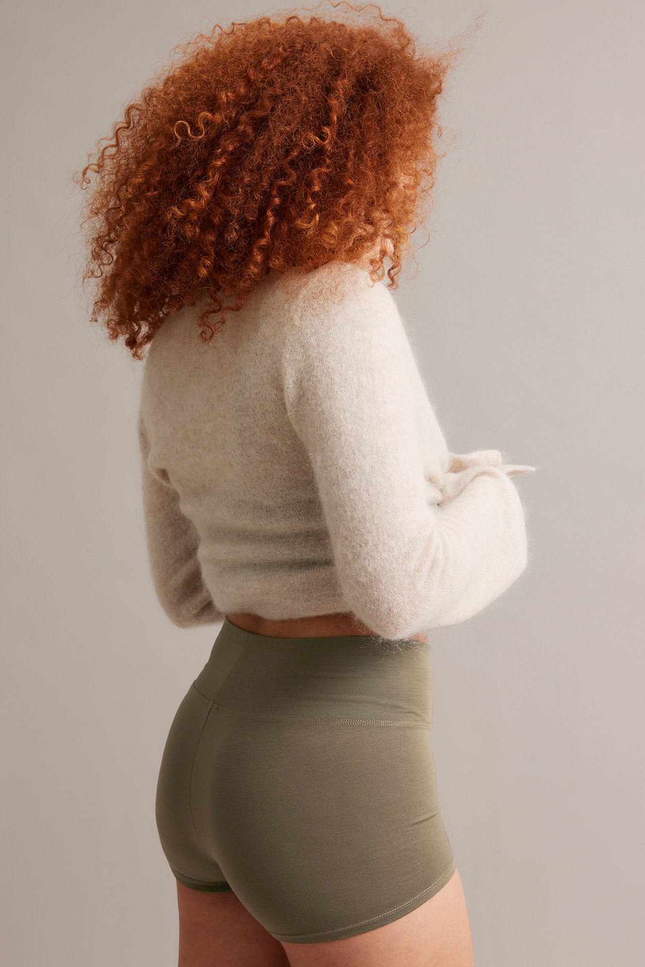 Sido-/ryggvy av kvinna i olivgröna, stretchiga boxertrosor och en ljusbeige fluffig kort tröja.