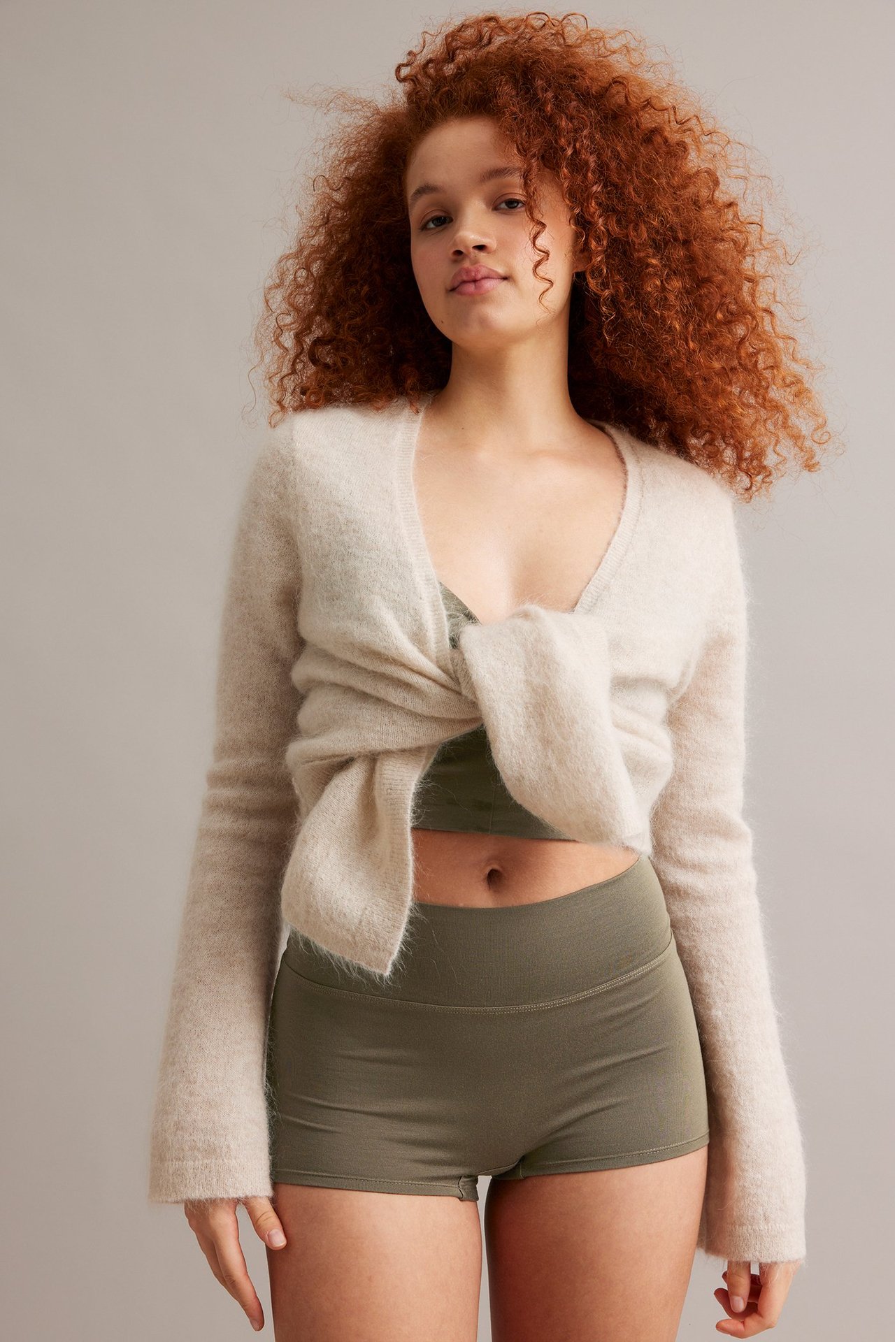 Forfra, kvinne i formtilpassede olivengrønne boksershorts, med matchende bralette og knyttet lys beige fluffy cardigan.