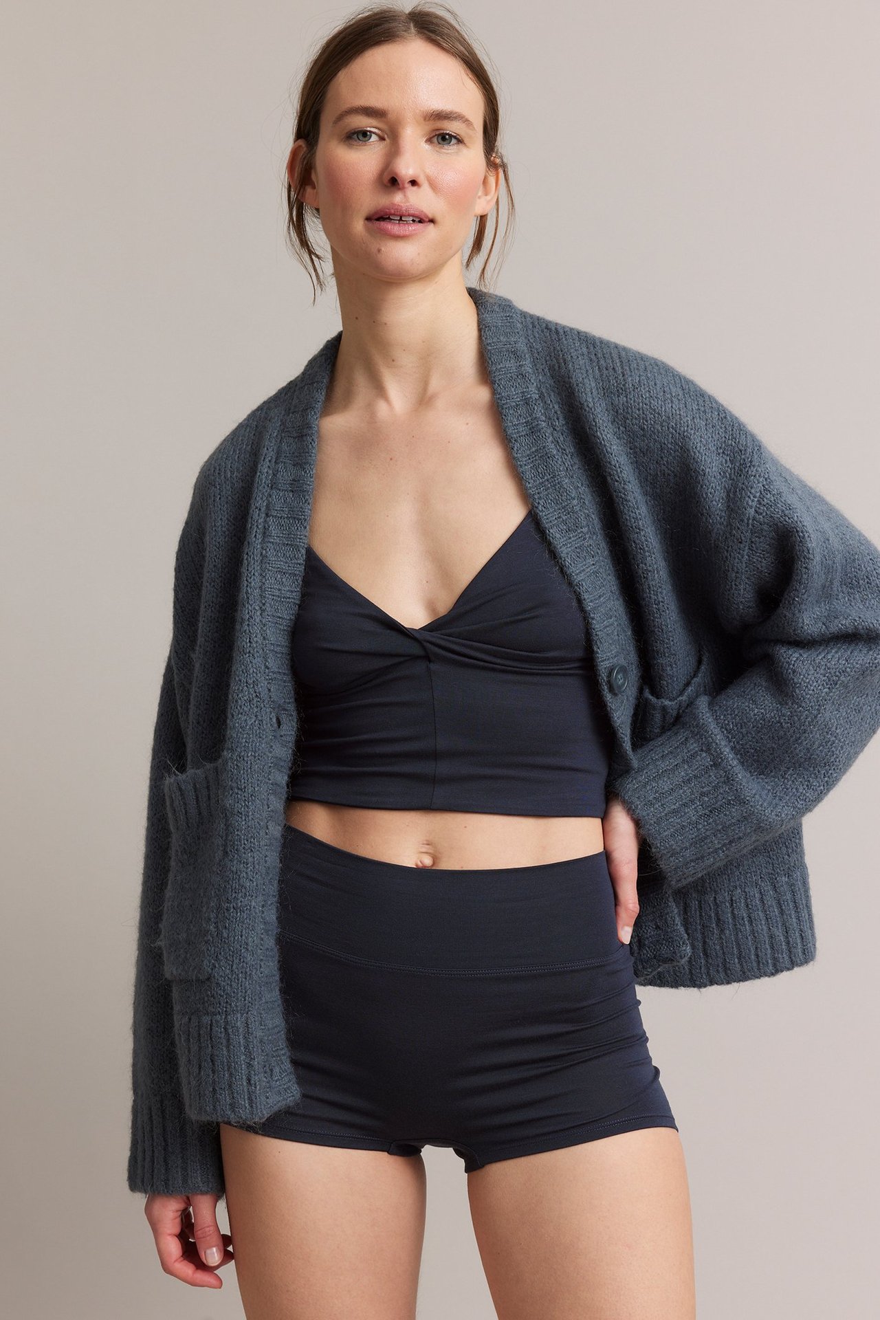 Frontvy av en kvinna i mörkblå boxertrosa i viskosblandning, matchande bralette och öppen gråblå stickad kofta.