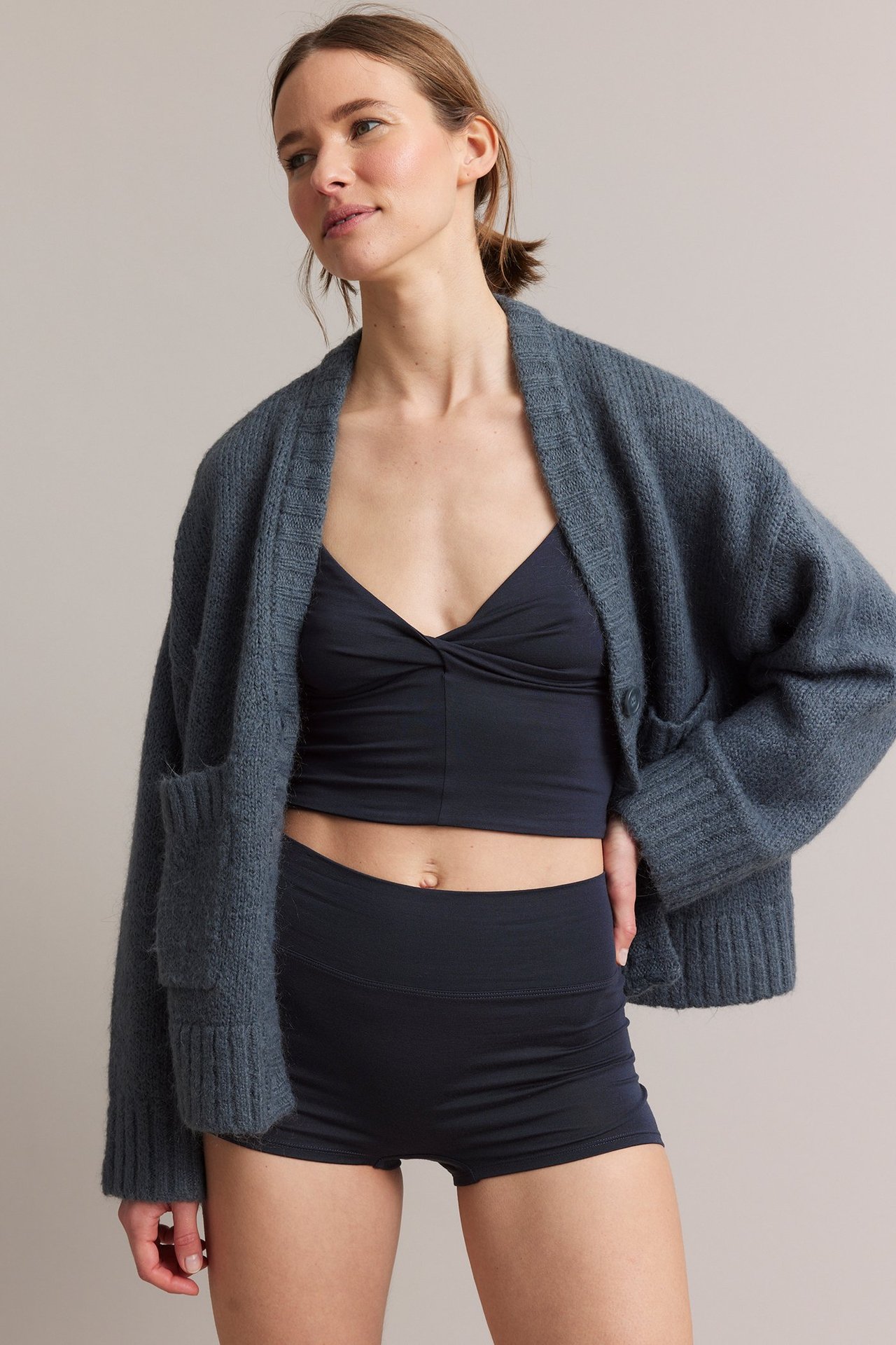 Frontvy av kvinna i mörkblå boxertrosa, matchande vriden crop top och gråblå stickad kofta.