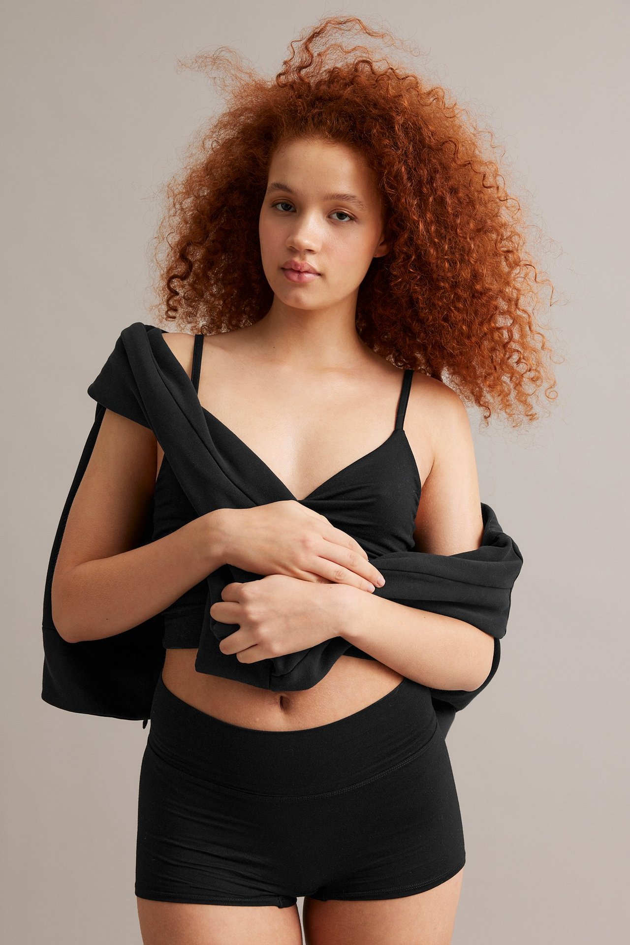 Forfra en kvinne i svarte boxershorts, en svart bralette og en svart topp drapert.