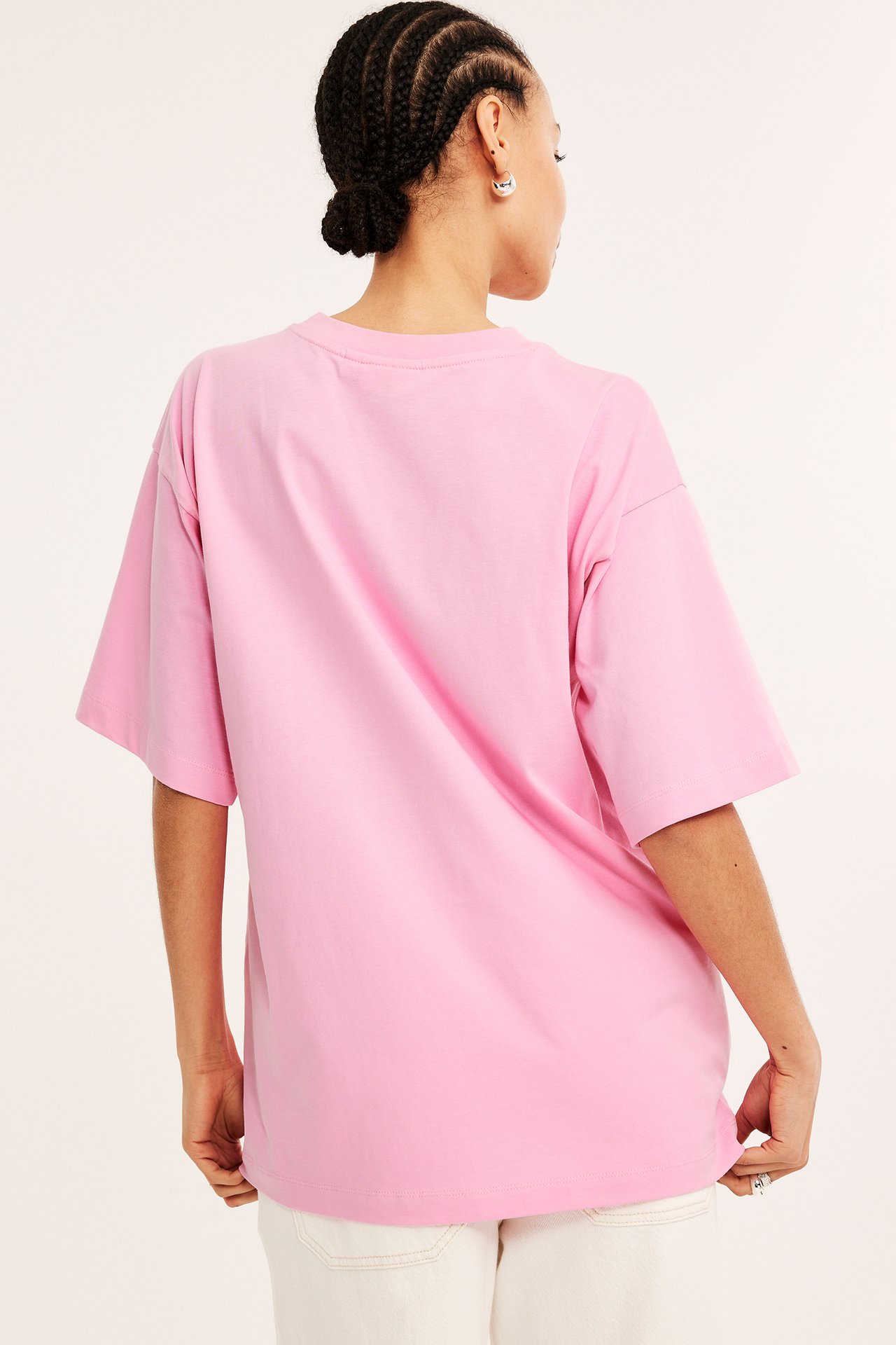 Bakifrån, kvinna i en ljusrosa oversized T-shirt i bomull, ljusa byxor och silverörhängen.