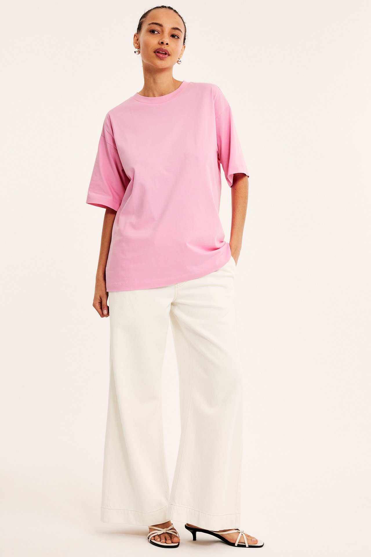 Framifrån: kvinna i ljusrosa oversized bomulls-t-shirt, vita vida byxor och vita remsandaler.